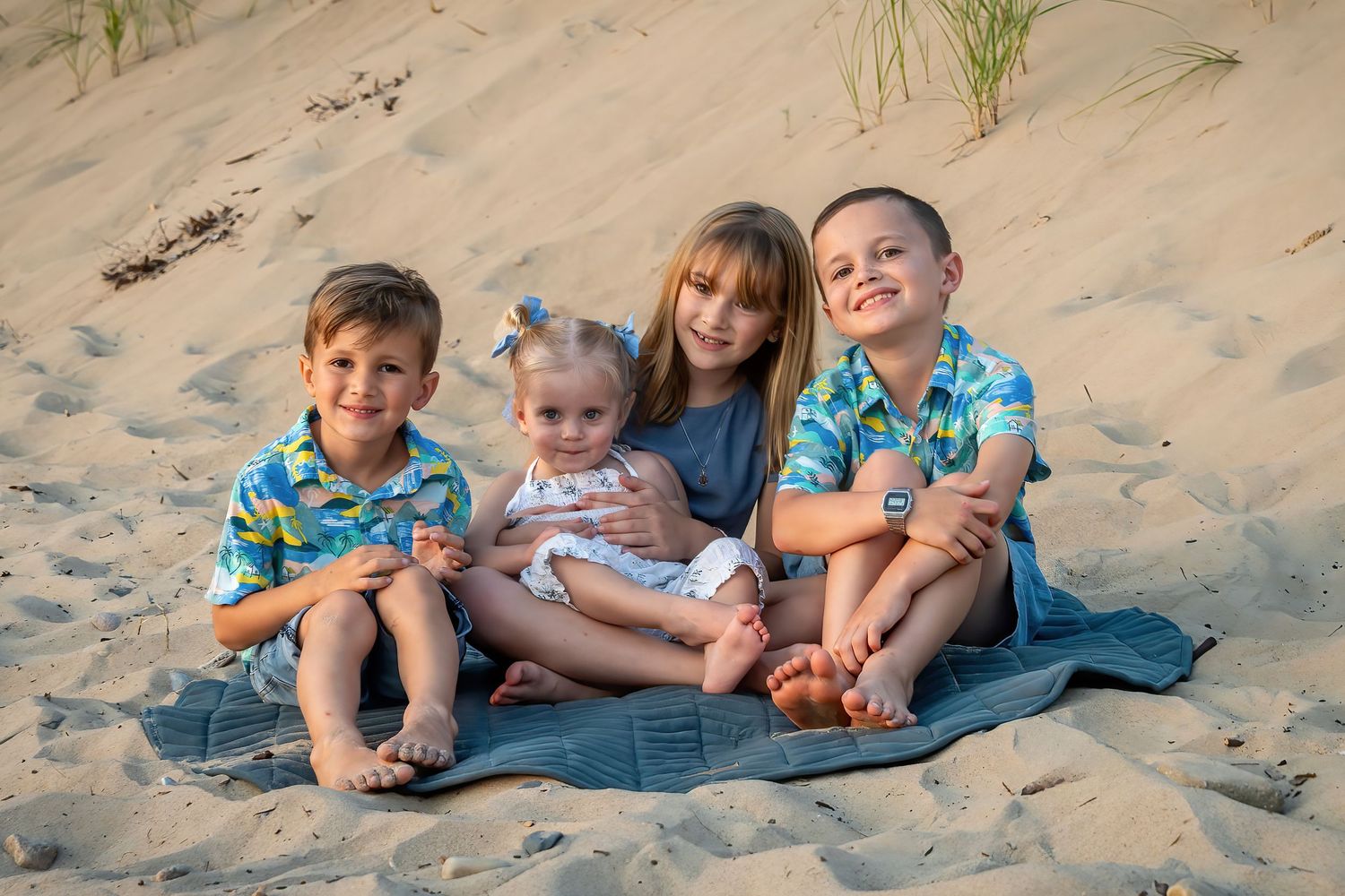 Four young children sitting together on sandy beach dunes smiling.