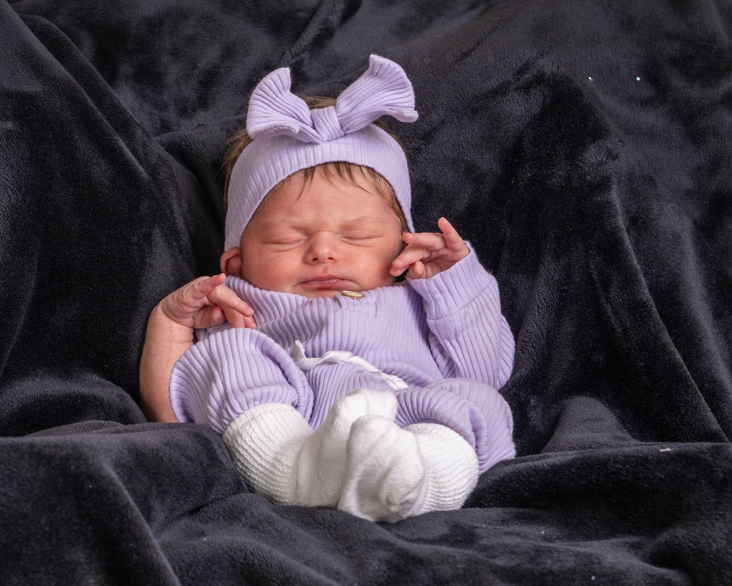 Peaceful newborn baby sleeping in lavender outfit and bow headband on dark velvet background.