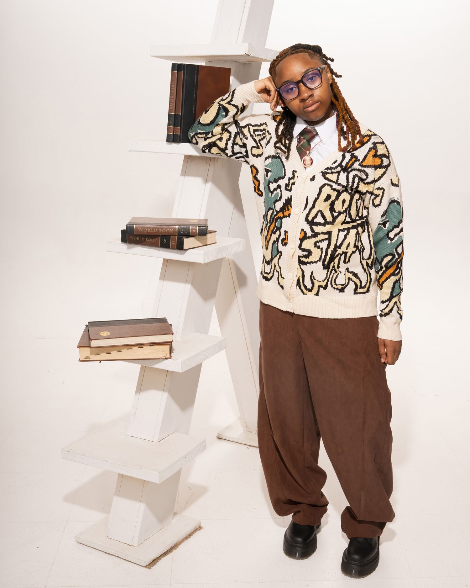 Young person leaning against white ladder bookshelf wearing colorful graphic cardigan and brown pants.