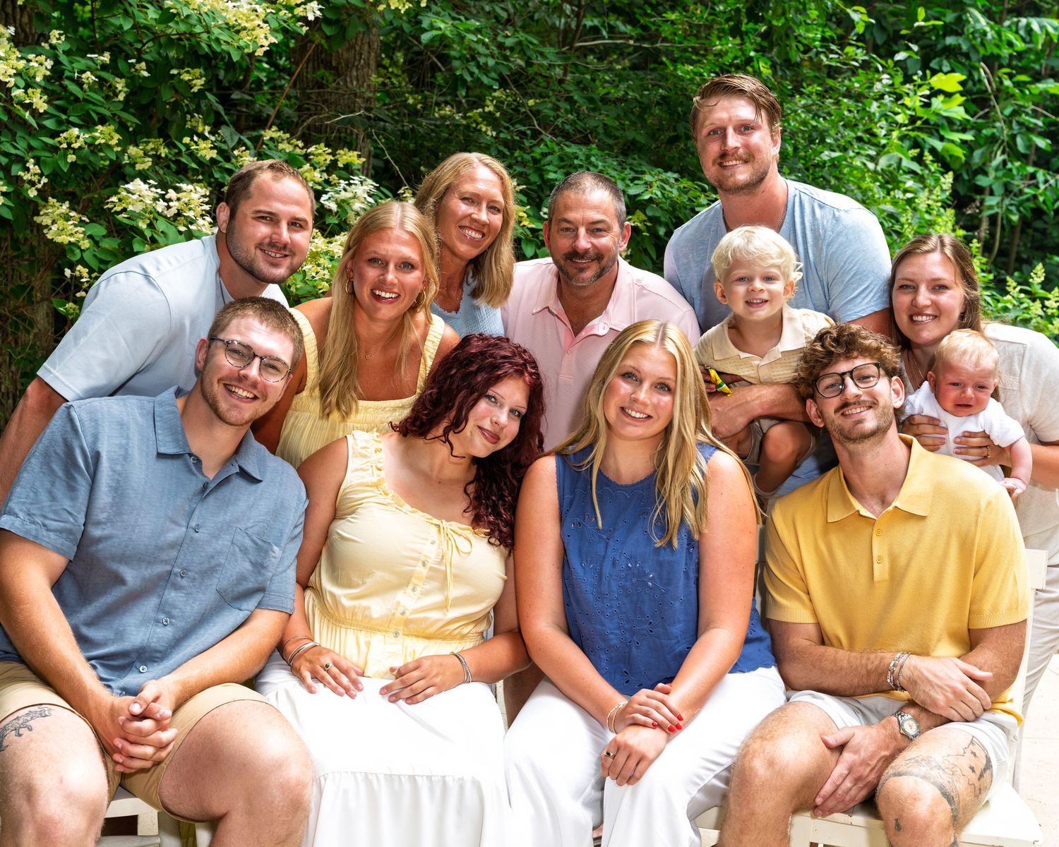 Large extended family group portrait smiling together outdoors surrounded by green foliage.