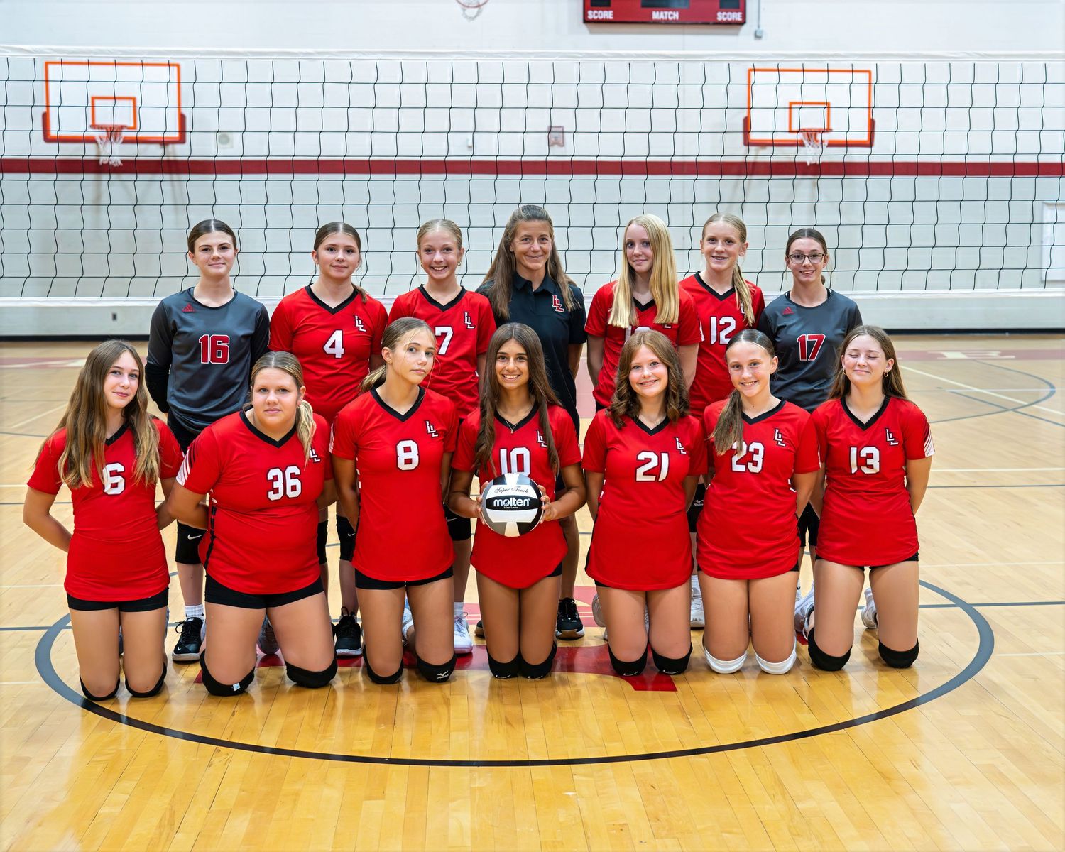 Girls volleyball team in red uniforms poses for official team photo in gymnasium with coaches and players.