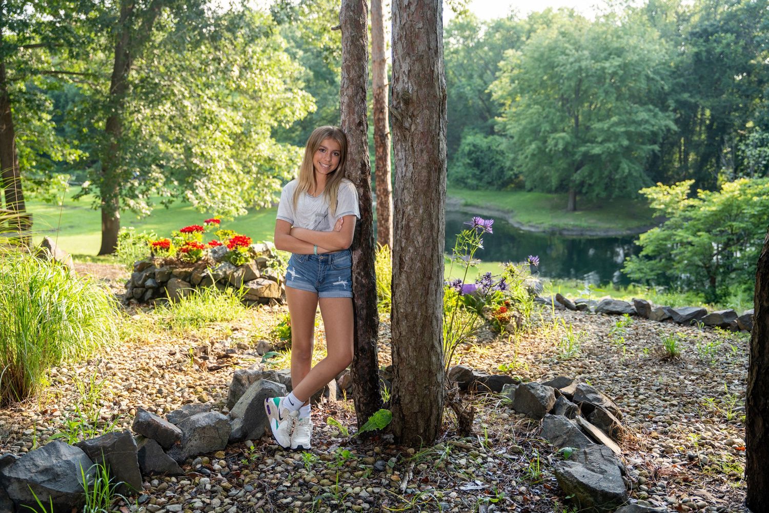 Young woman in denim shorts leaning against a tree trunk surrounded by lush greenery and a garden pond.