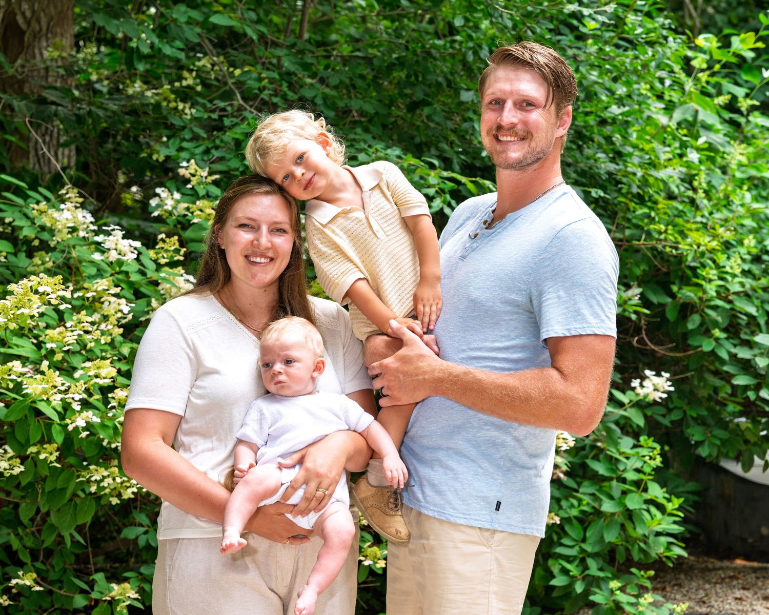 Happy young family of four with newborn baby and toddler posing in lush garden setting.