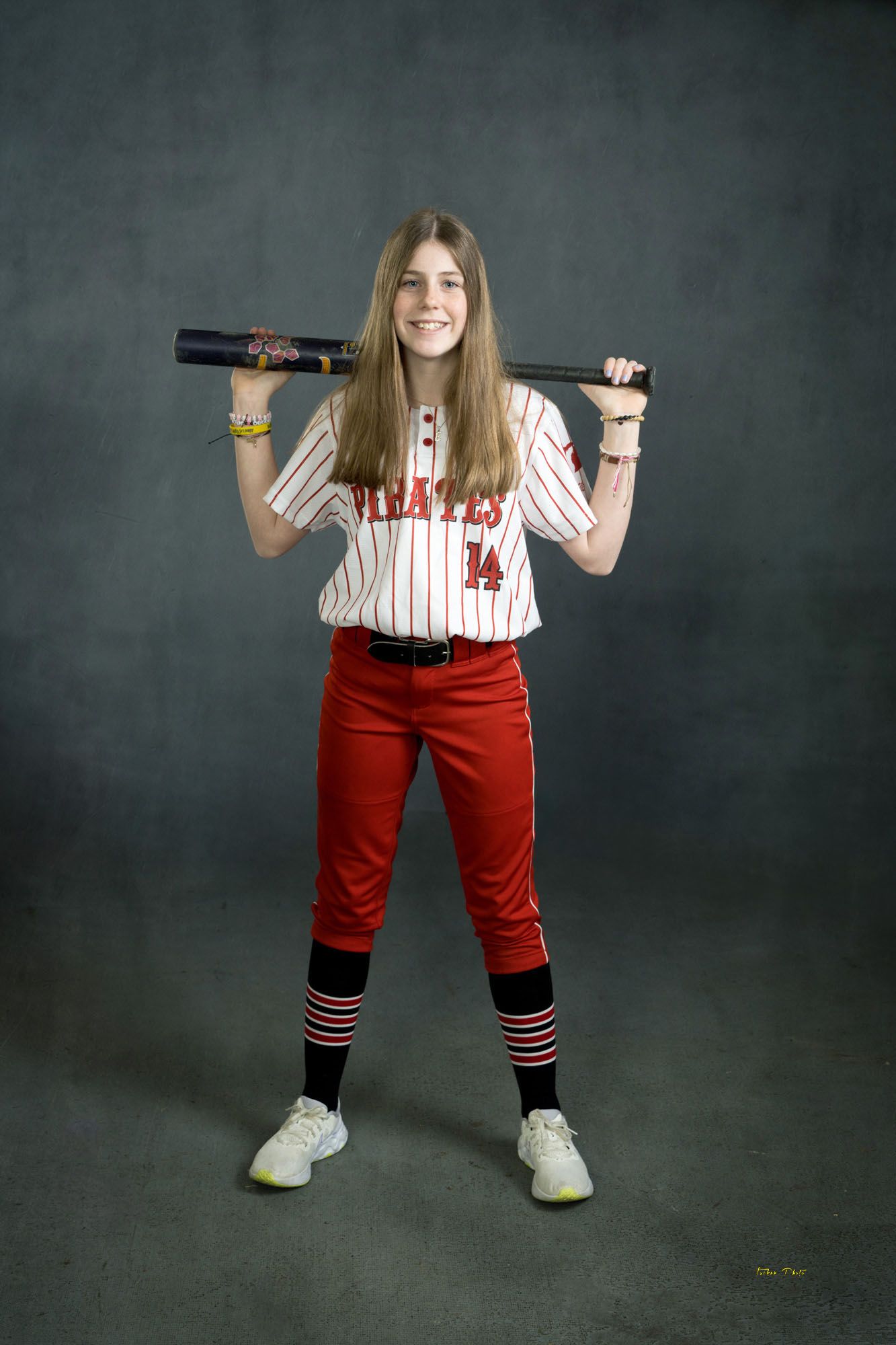 Smiling female Pirates softball player #14 poses with bat across shoulders in red and white pinstripe uniform.