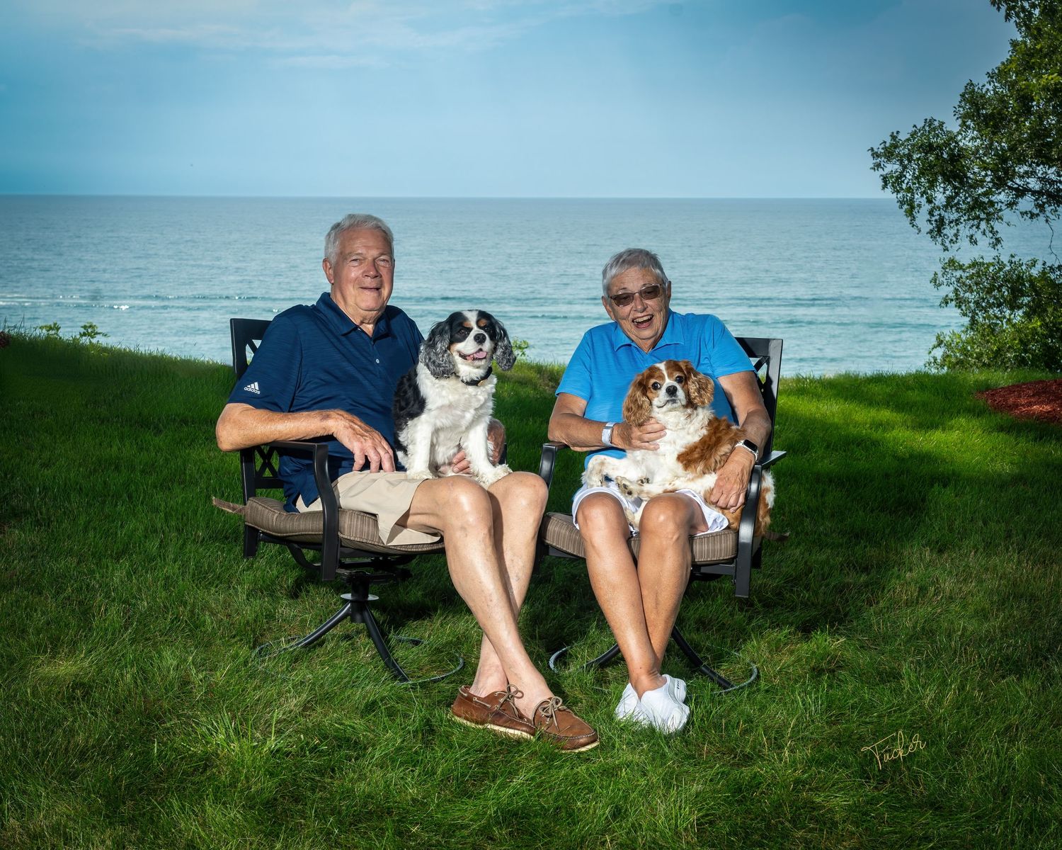 Two elderly people sitting in chairs on grass near lake, each holding a small dog on their laps.