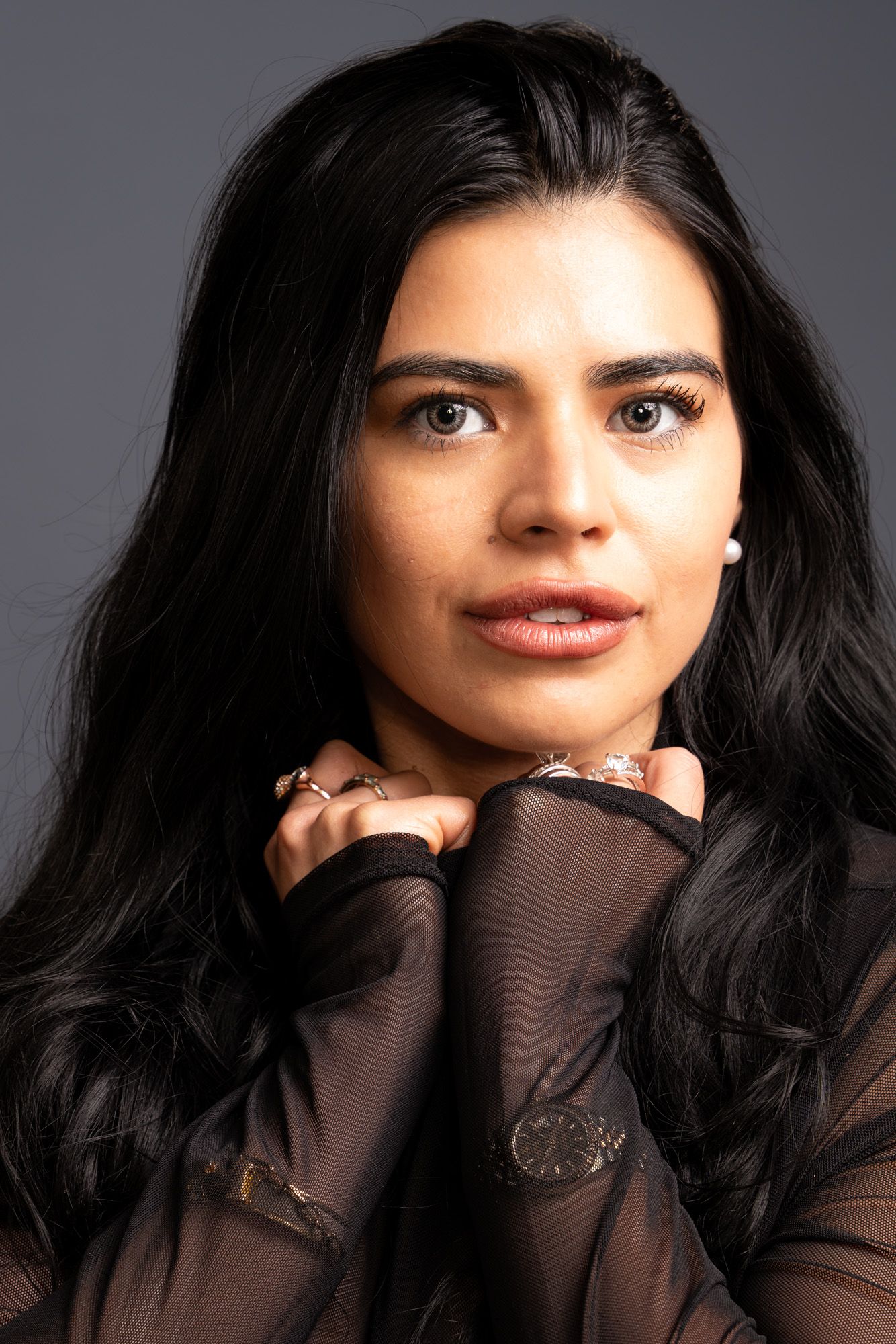 Beautiful dark-haired woman in sheer black top with rings and watch looking away softly.