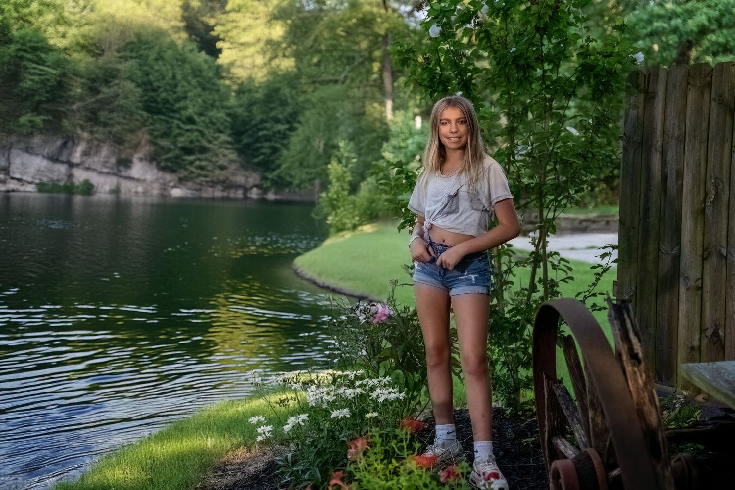 Young blonde woman in denim shorts and white tee poses by a serene lake surrounded by lush green trees and colorful flowers.
