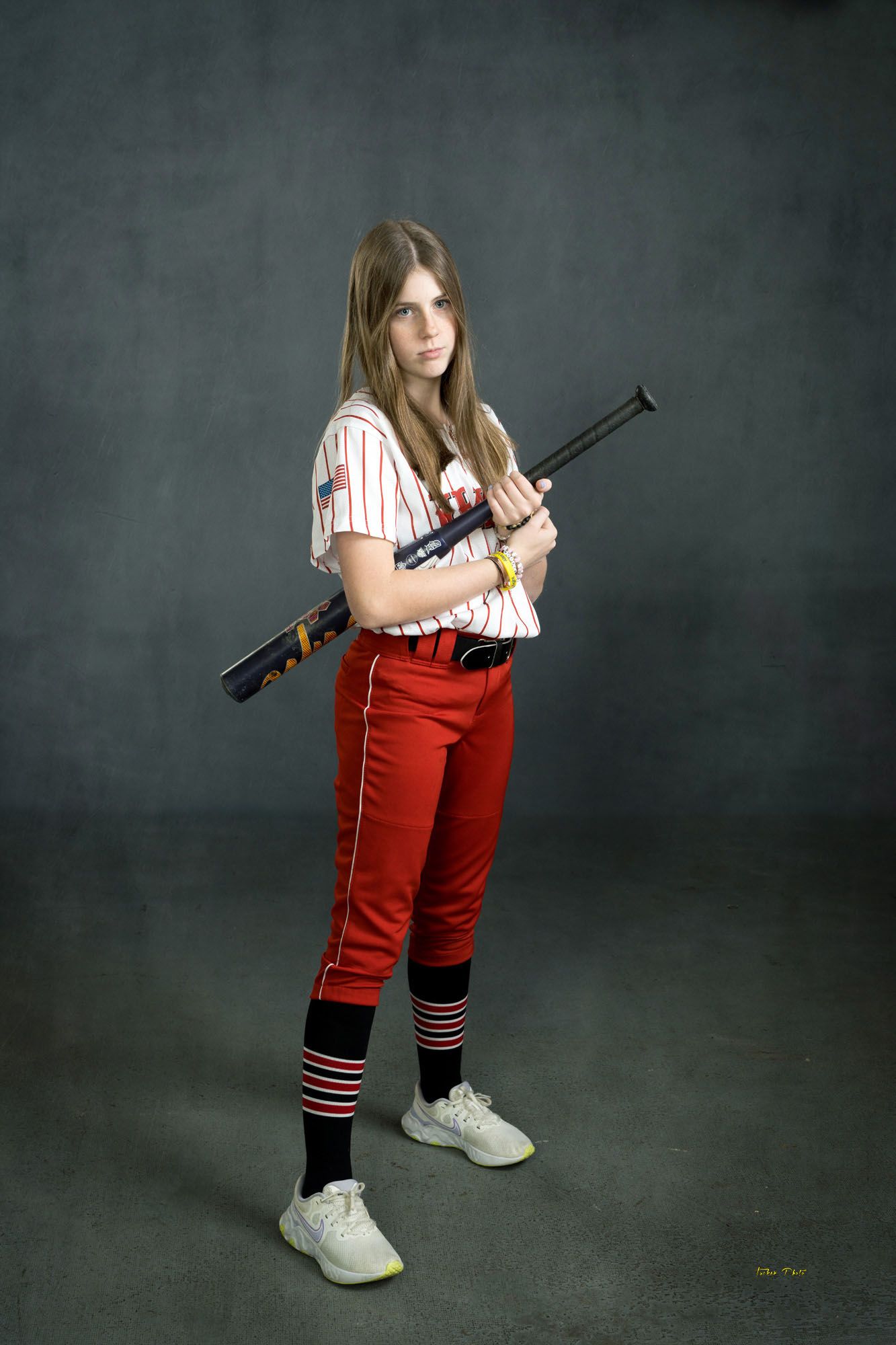 Serious female softball player in red and white uniform posing with black bat on shoulder.