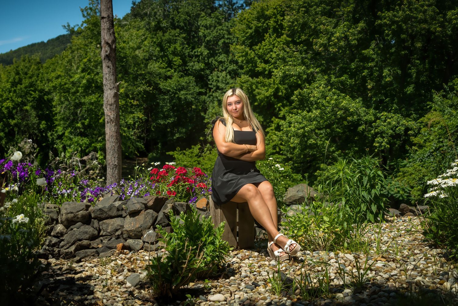 Blonde woman in black dress sitting among colorful garden flowers with lush green trees in background.
