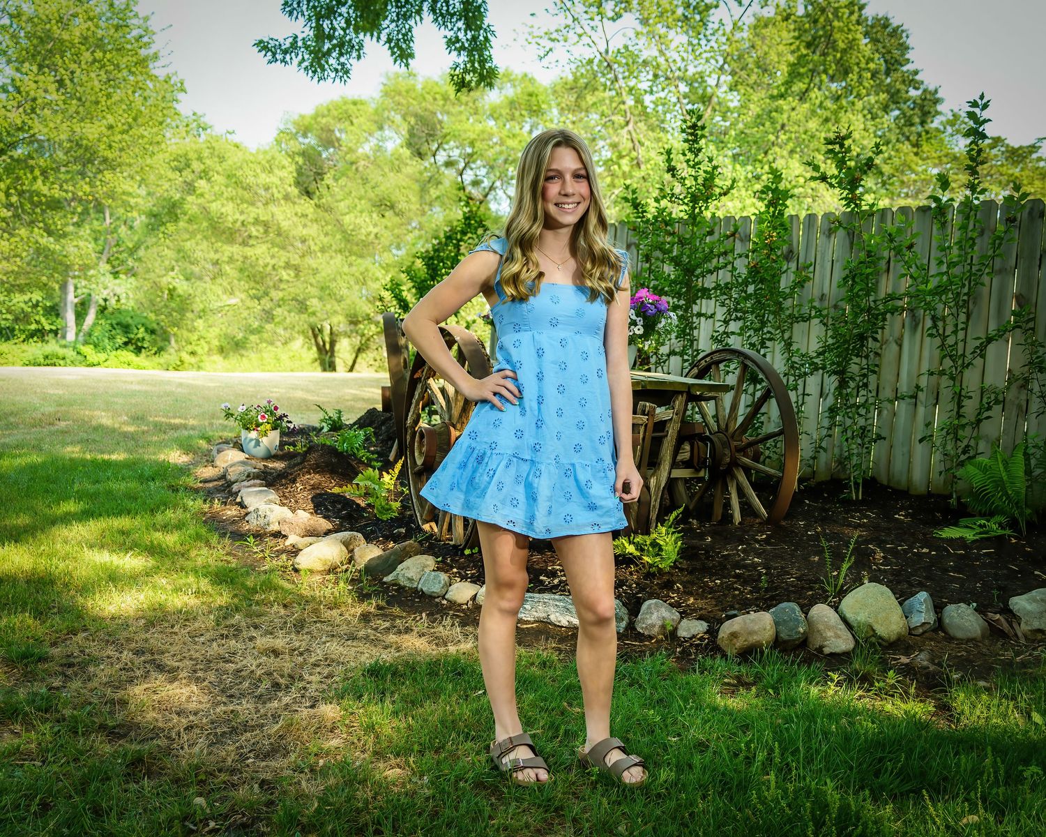 Smiling young woman in blue spaghetti strap dress standing on green lawn with garden and wagon wheel.