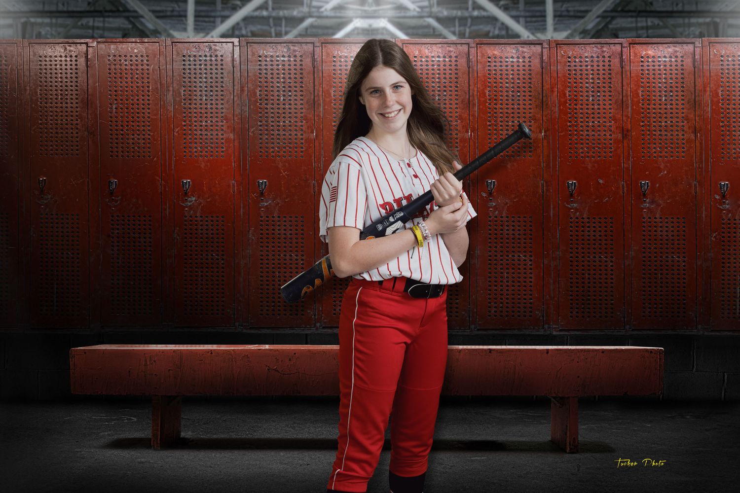 Smiling girl in baseball uniform holding a bat in front of red lockers wearing red and white uniform.