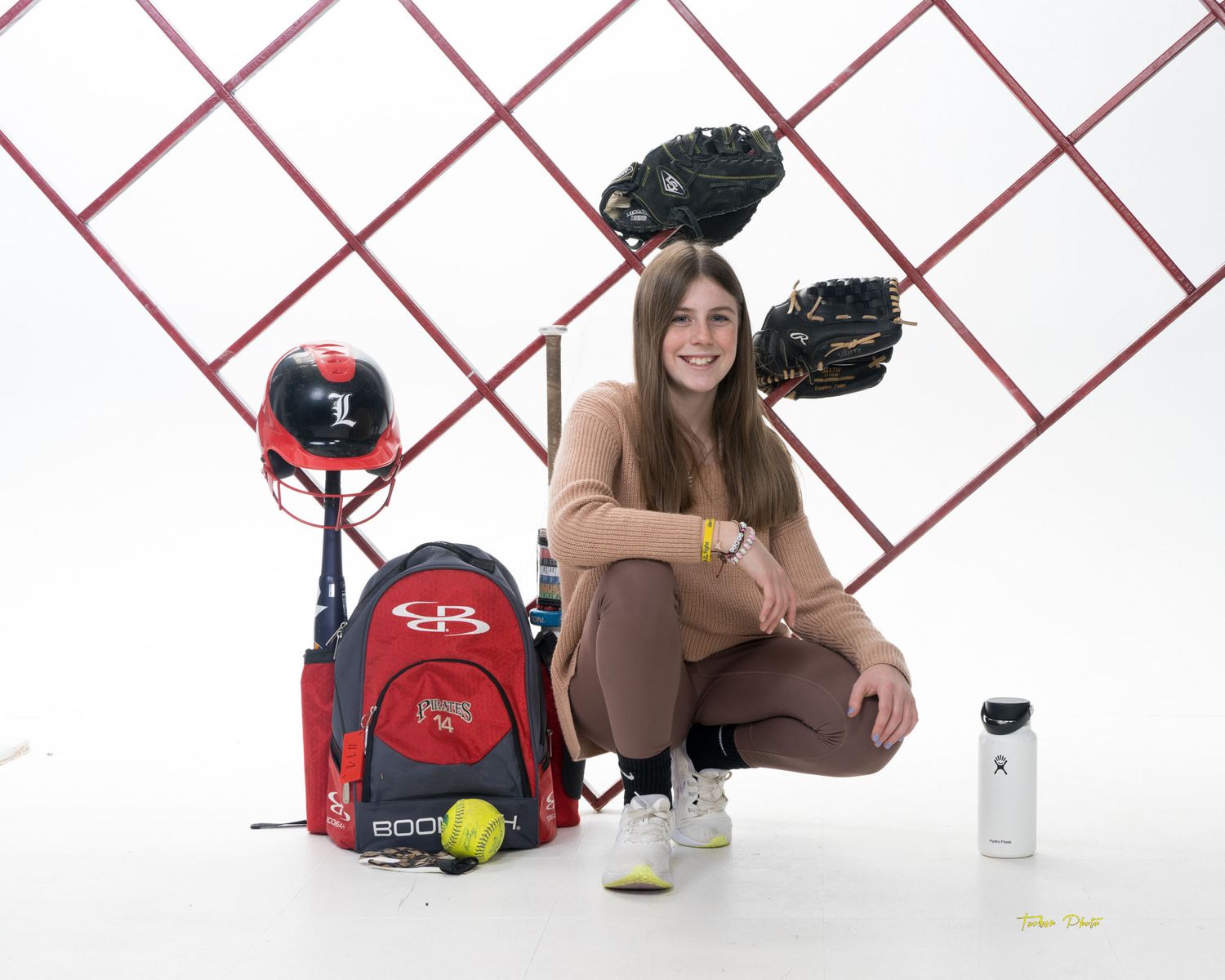 Female softball player crouches beside her Pirates softball equipment including backpack, helmet, and gloves in a studio.