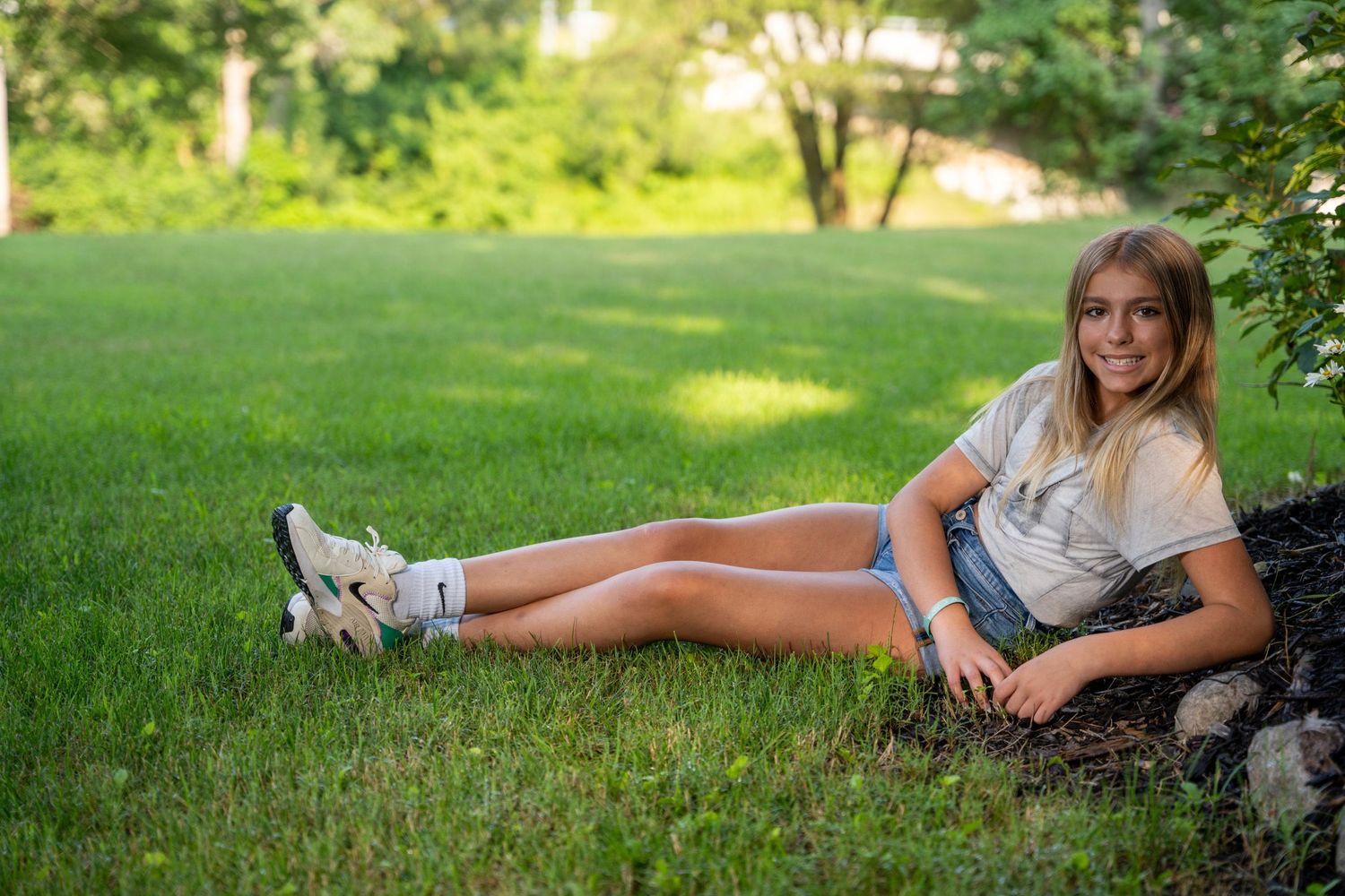 Smiling teenage girl in denim shorts sitting on green grass leaning against a tree in a sunny park.