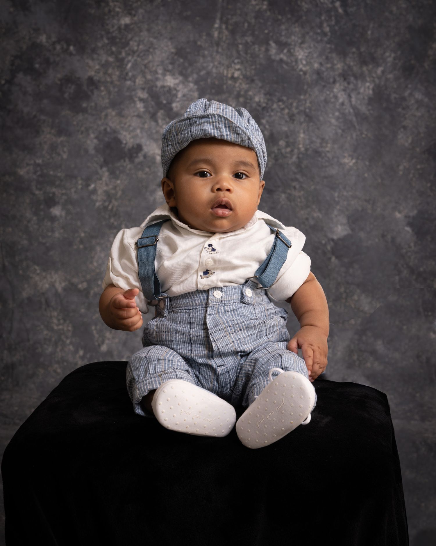 Adorable baby boy in blue suspender outfit and cap seated on dark surface in studio.