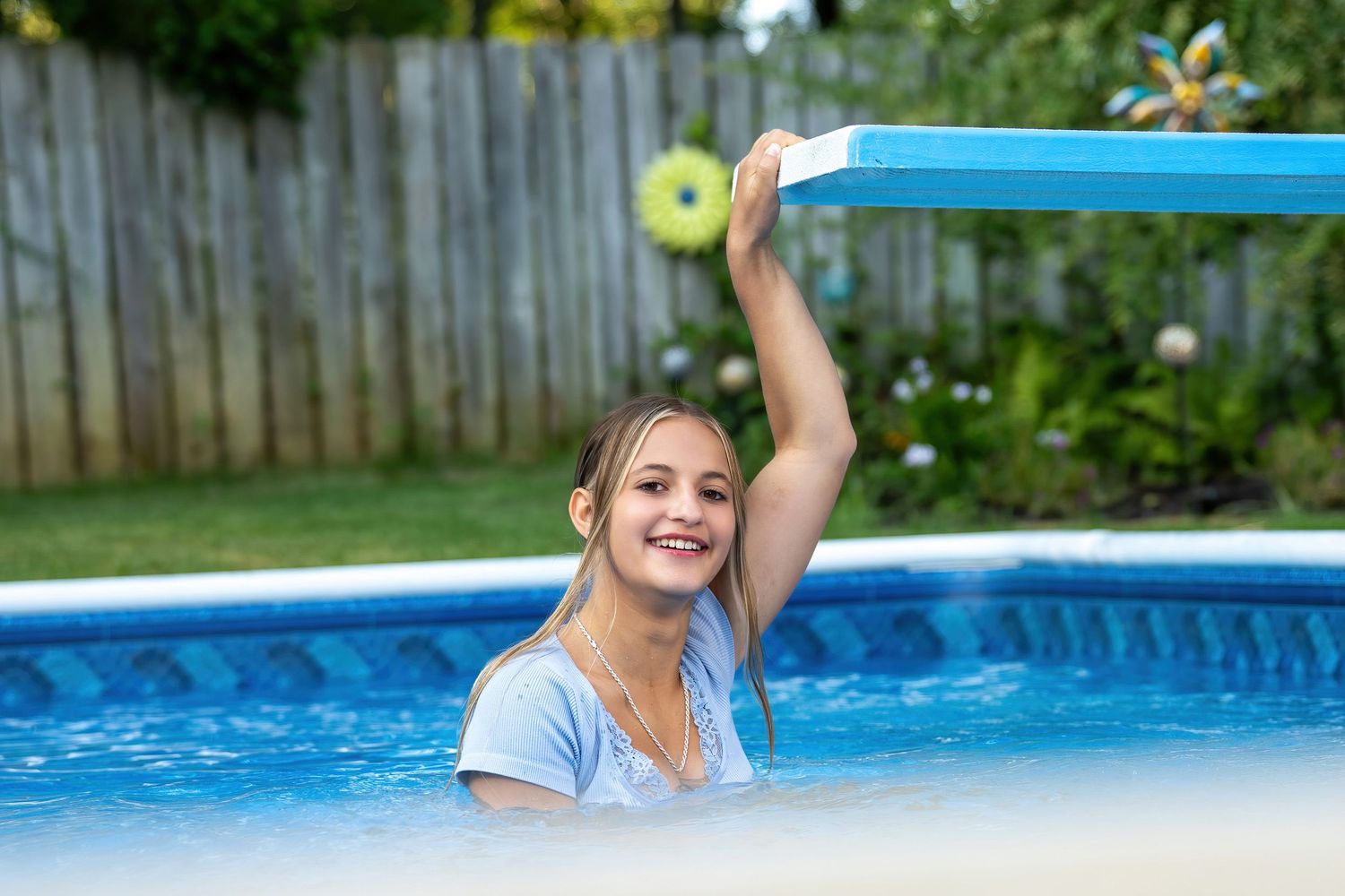 Smiling girl in a pool holding a blue pool noodle overhead on a sunny day.