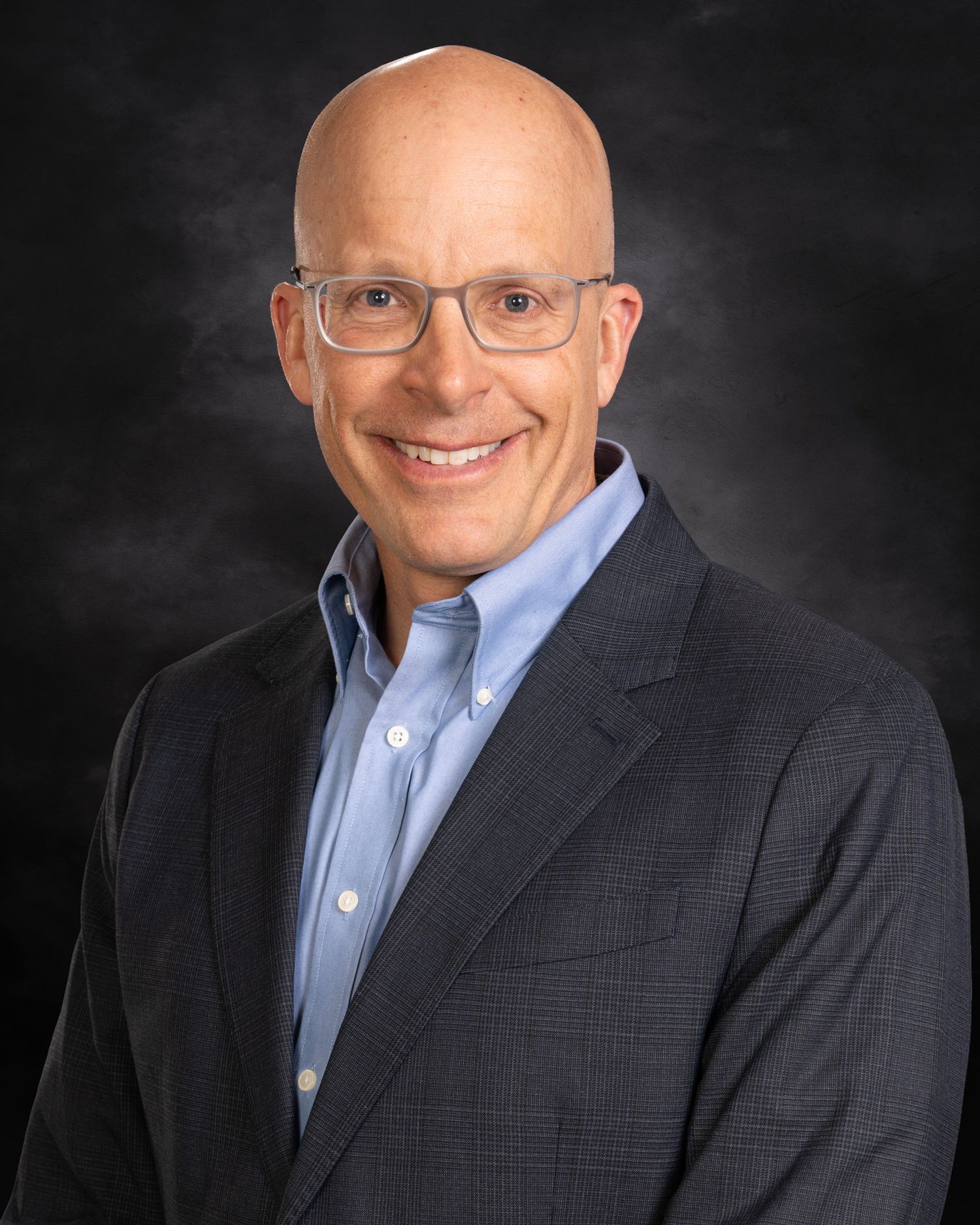 Business headshot of a bald man in glasses and navy blazer smiling confidently against a dark studio background.
