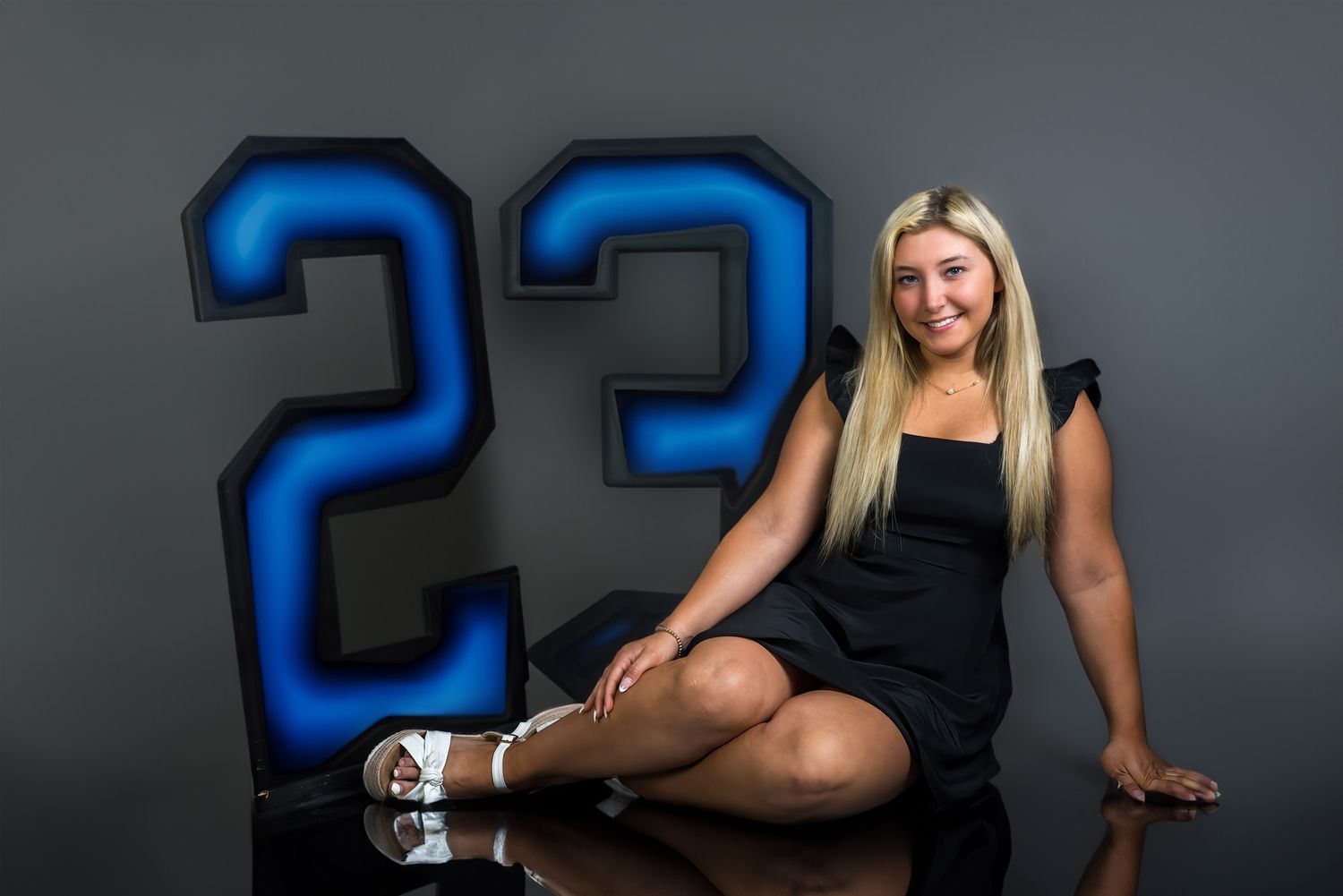 Smiling blonde teen in black dress posing beside illuminated number 23 sign for senior portrait.