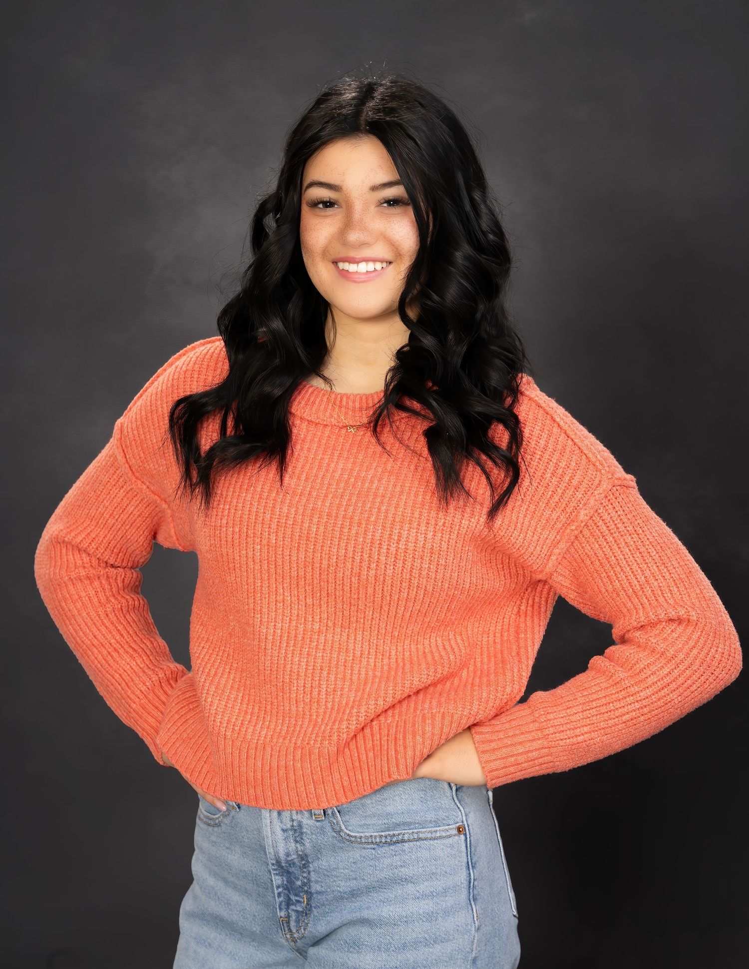 Smiling young woman in an orange ribbed knit sweater and jeans posing with hands on hips on dark backdrop.