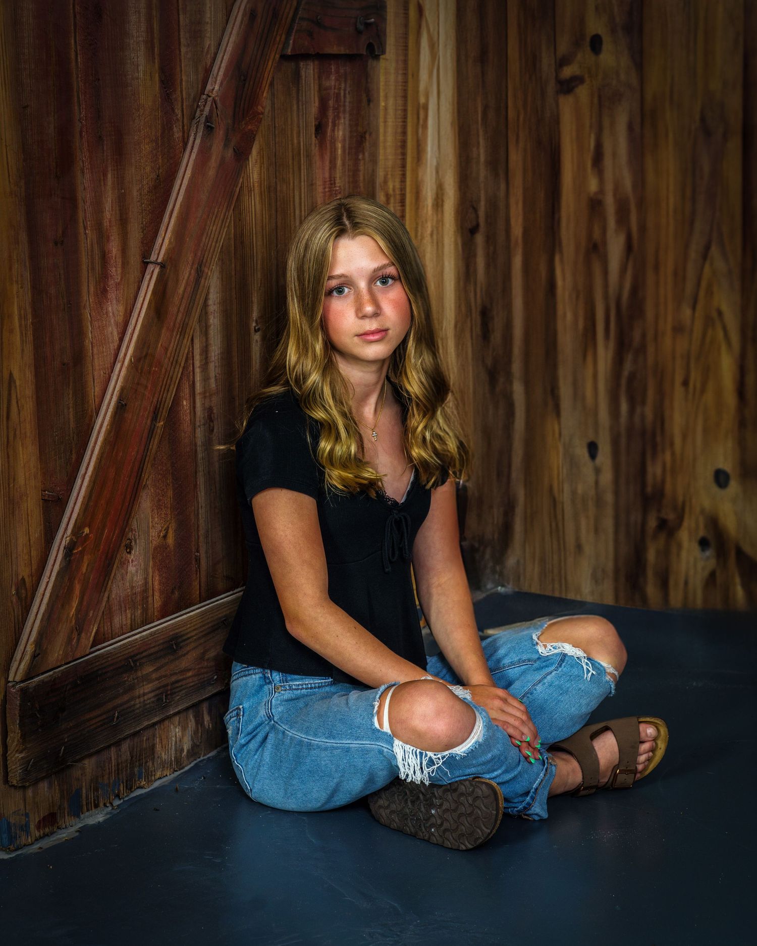 Teen girl in black top and ripped jeans sitting on floor leaning against a rustic wooden barn door.