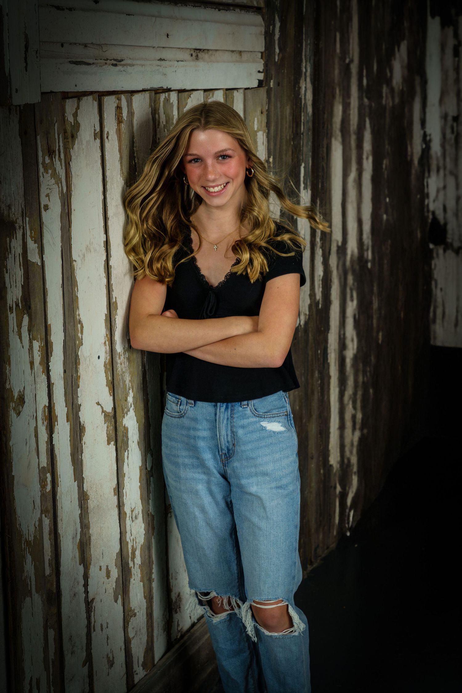 Smiling teenage girl in black top and ripped jeans standing with arms crossed against a weathered white barn wall.