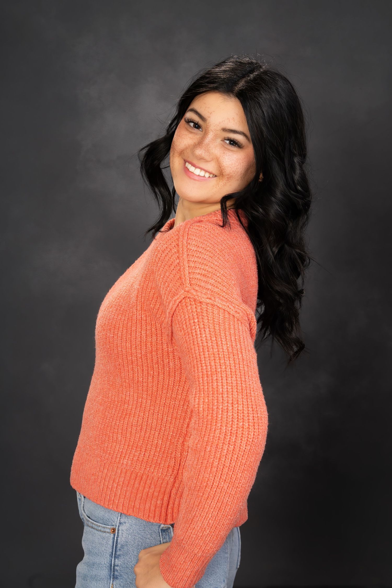 Young woman smiling in coral knit sweater and jeans against dark background.