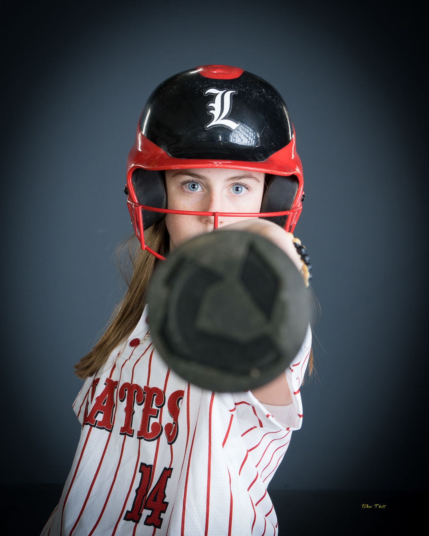 Young softball player in Pirates uniform wearing red helmet pointing bat toward camera.