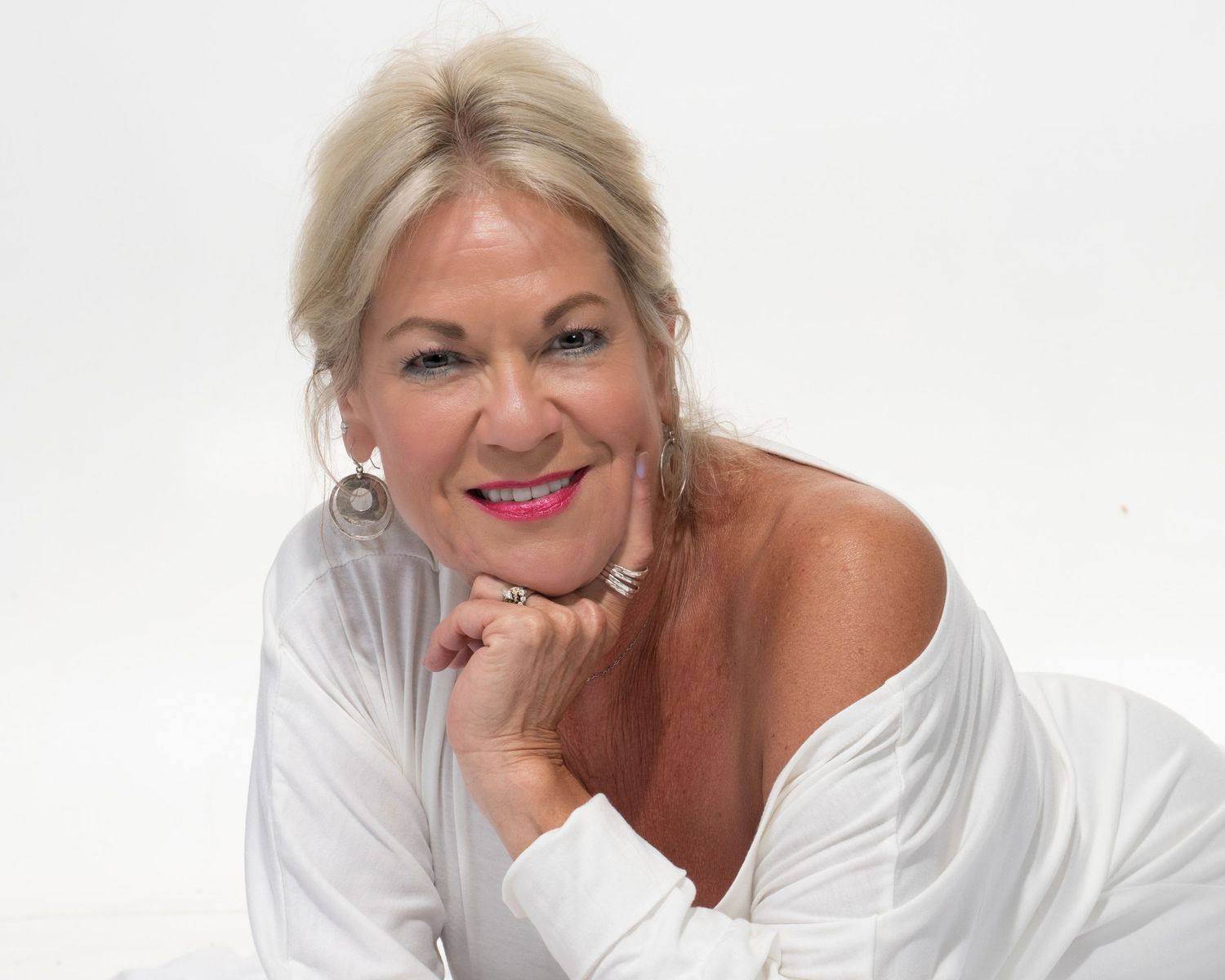 Blonde woman in white off-shoulder top posing on white background with chin on hand.