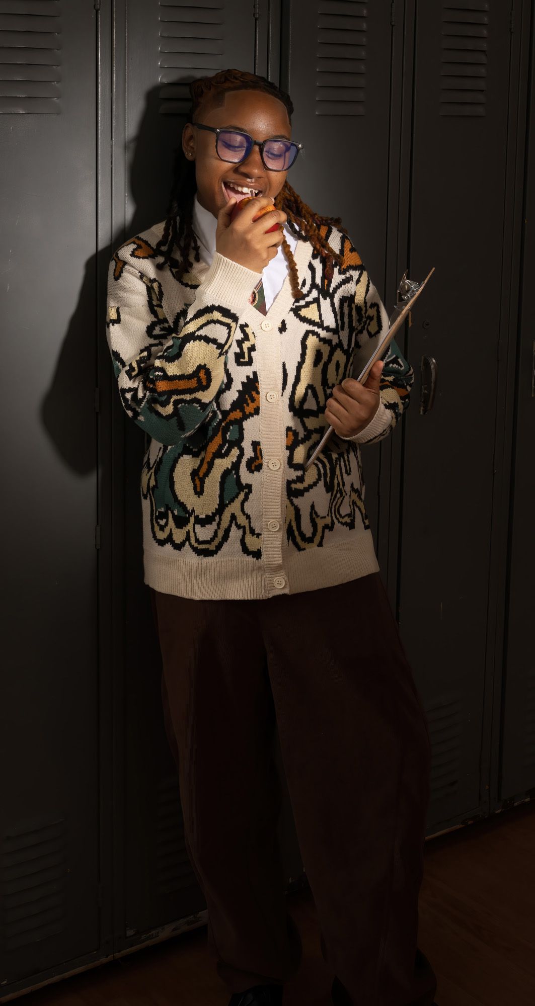 Smiling student in graphic cardigan holding clipboard by school lockers.