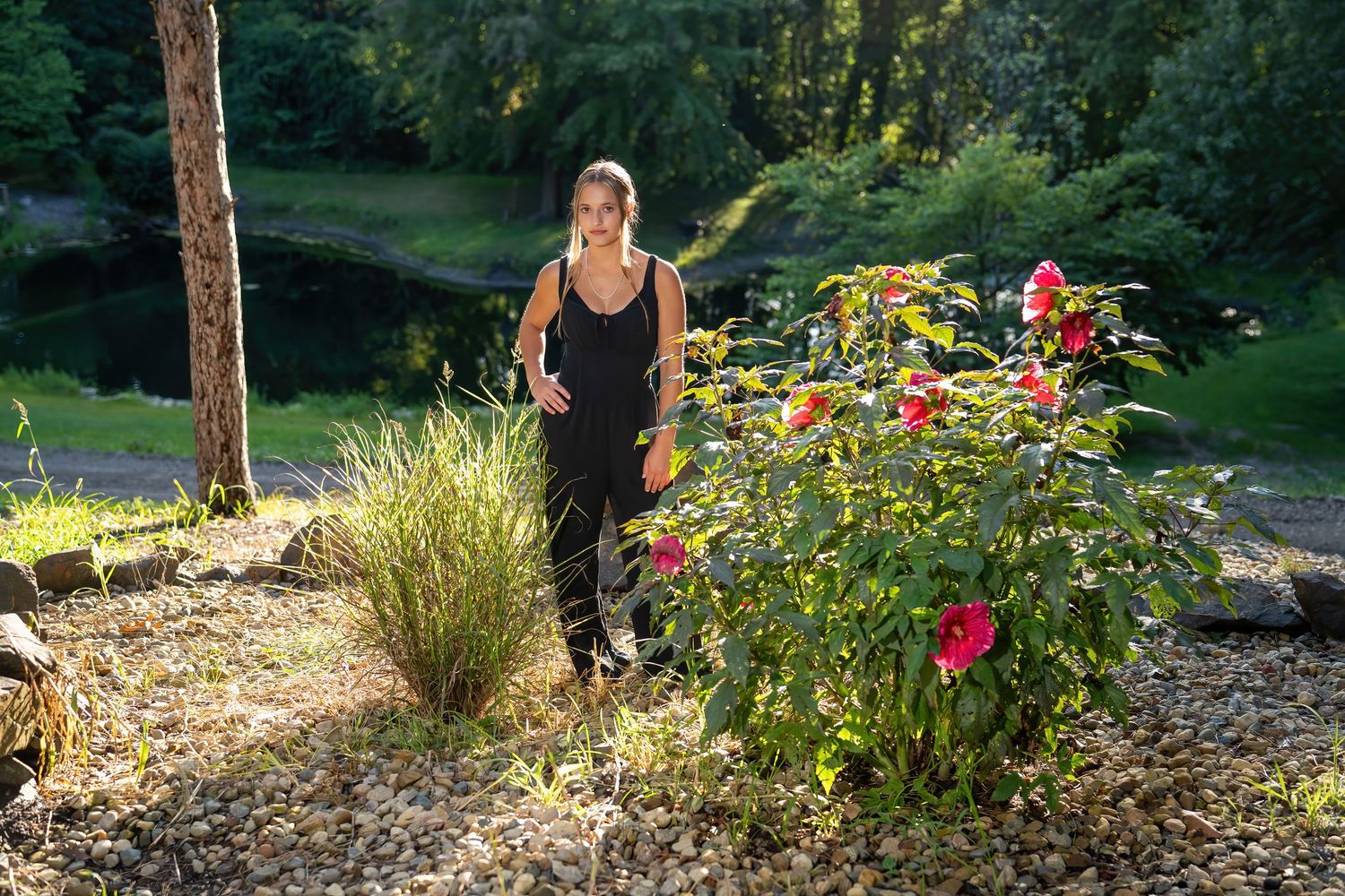 Woman in black sleeveless jumpsuit strolls past vibrant red roses in sunny outdoor garden setting.