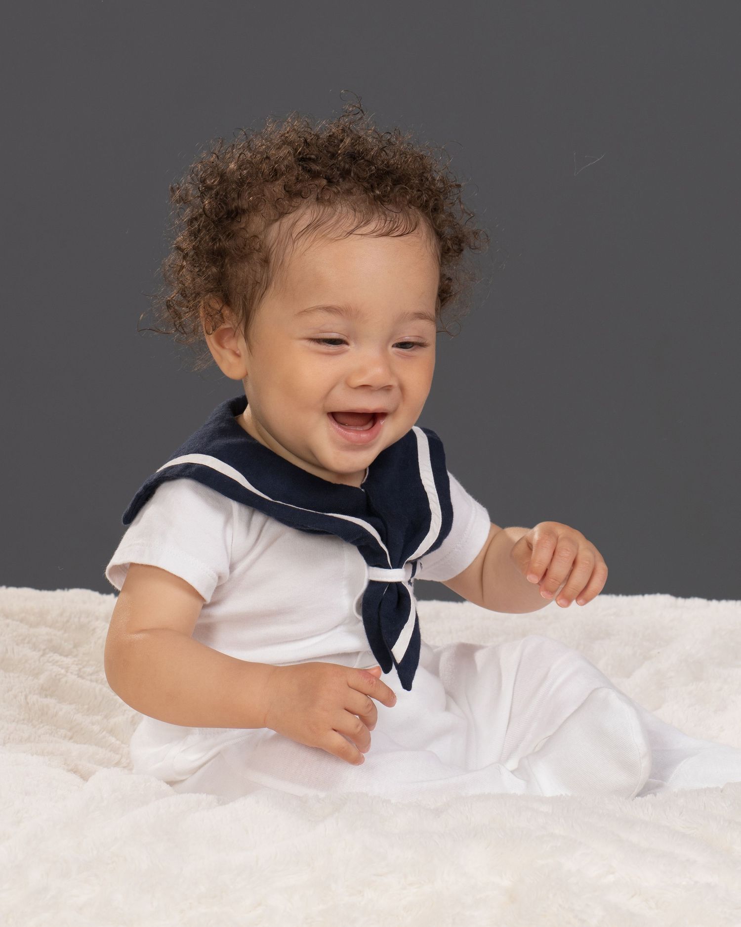 Happy toddler in white sailor outfit sitting on fluffy blanket against gray background.