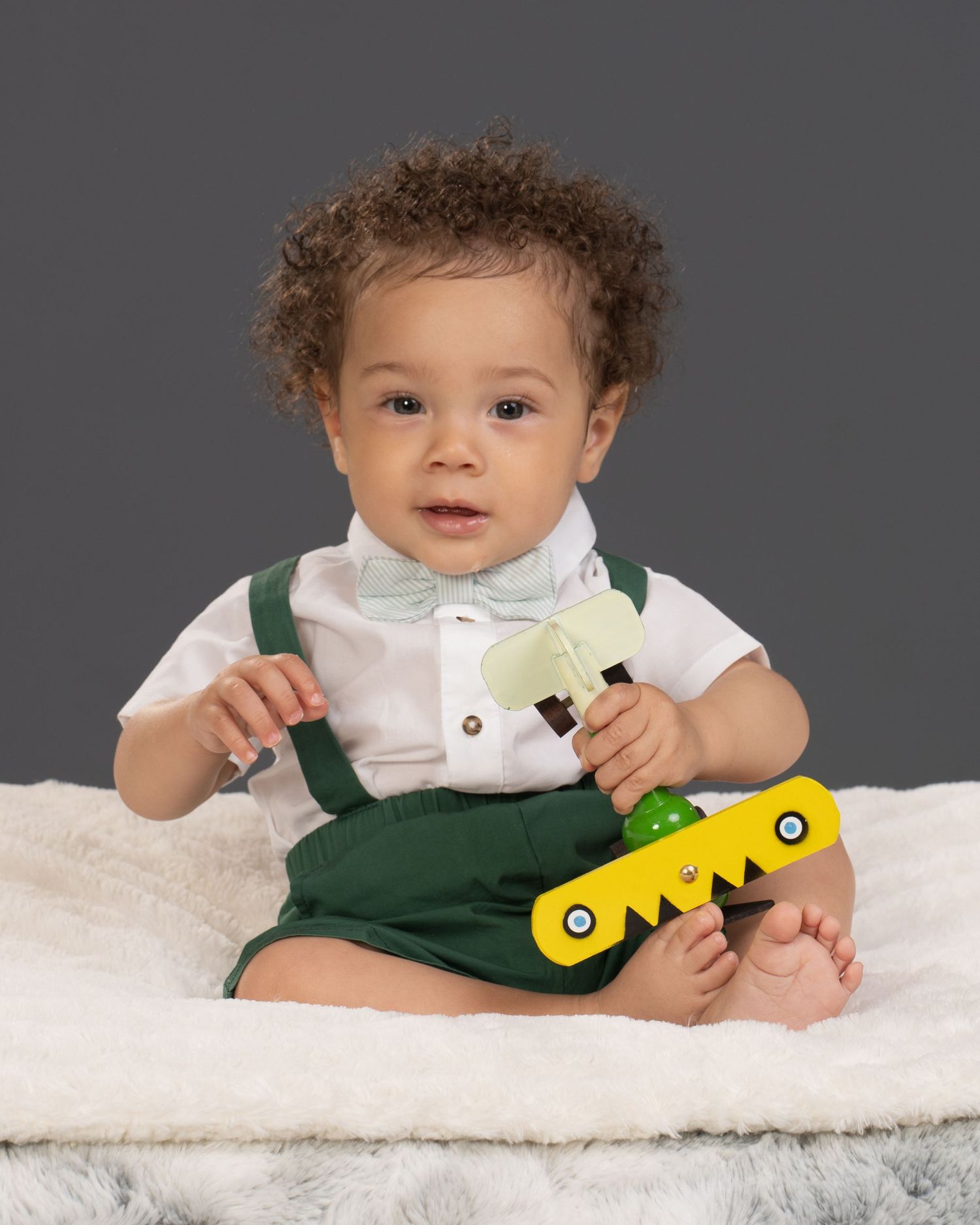 Cute baby boy in green overalls with bow tie playing with colorful toy on white blanket.