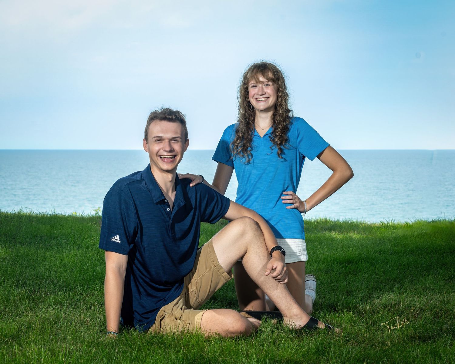 Teen boy and girl posing together on grassy hillside with lake and blue sky in background.