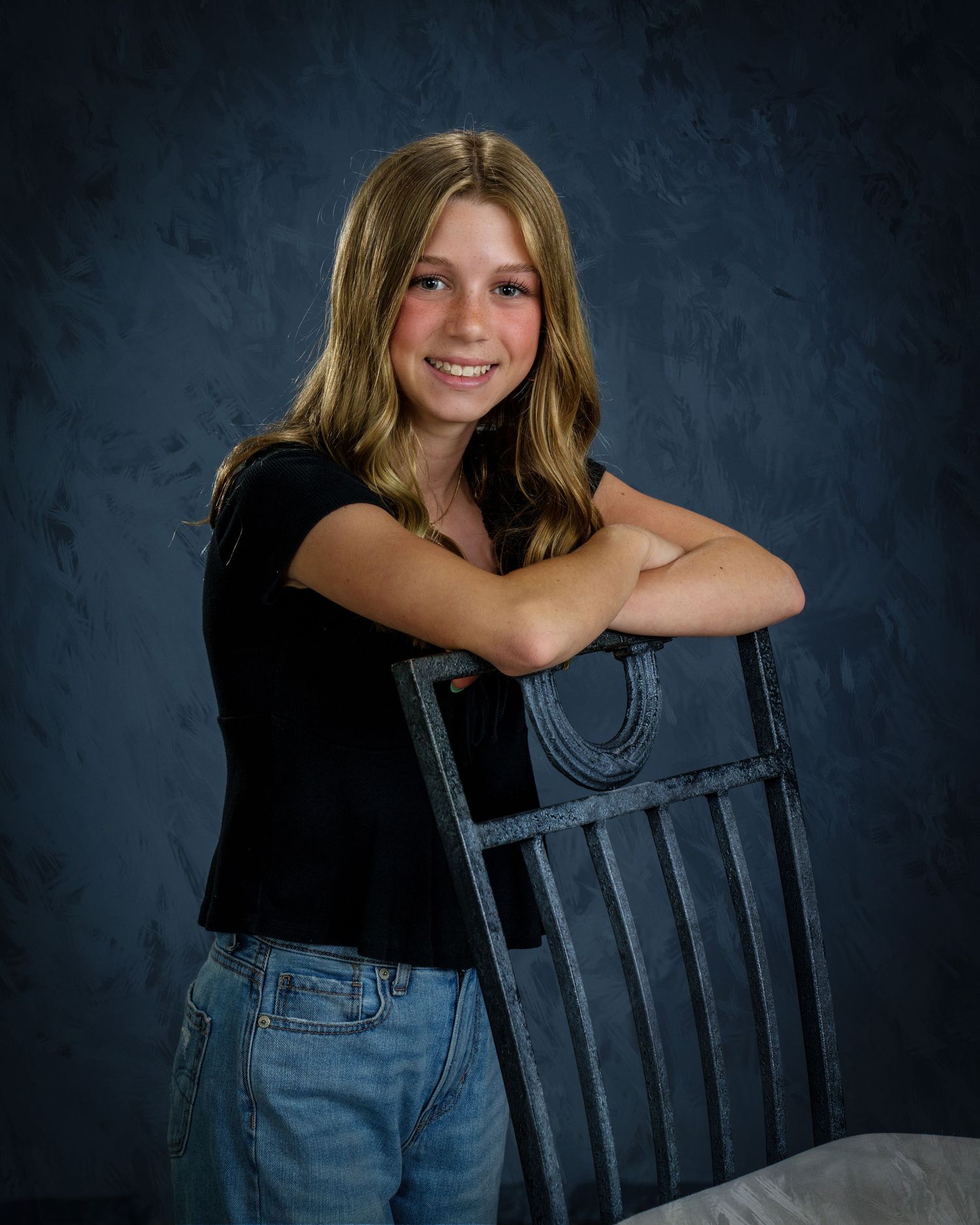 Smiling teen girl in black top and jeans leaning on a decorative chair against a blue textured backdrop.