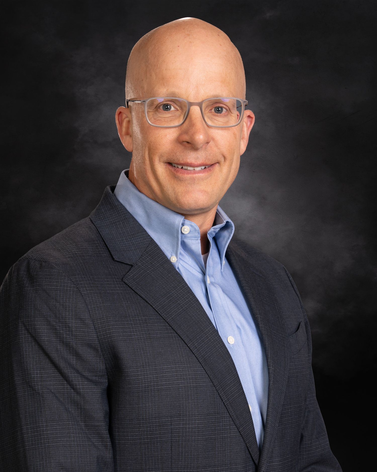Professional headshot of a bald man wearing glasses and a navy blazer over a light blue shirt against dark background.