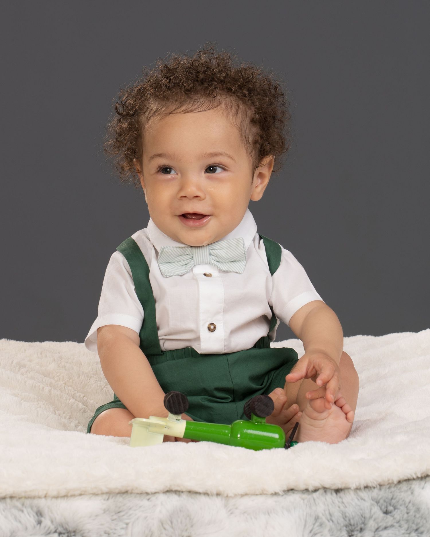 Happy smiling baby in green suspenders and white shirt with bow tie on fluffy blanket.