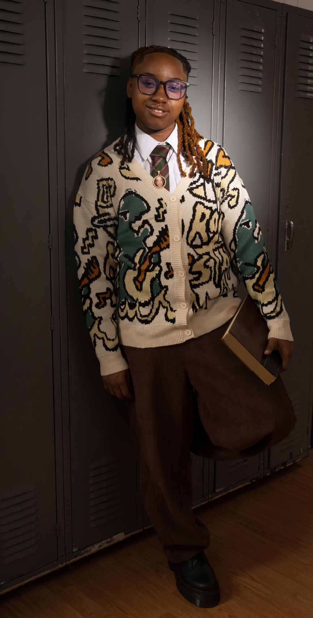 Stylish student in patterned cardigan holding book standing by gray lockers.