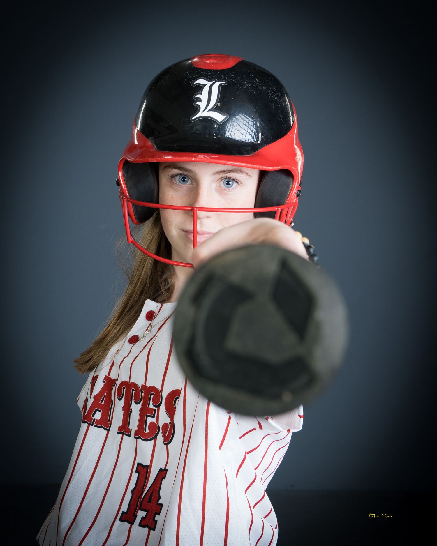Youth softball player number 14 in Pirates uniform with red helmet aiming bat forward.