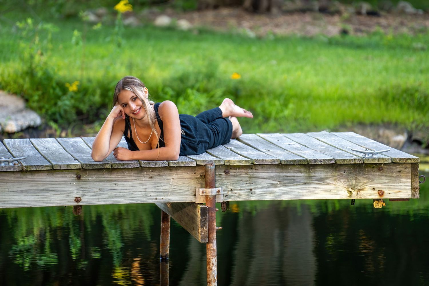 Young woman relaxes on wooden dock overlooking calm reflective pond surrounded by lush green landscape.