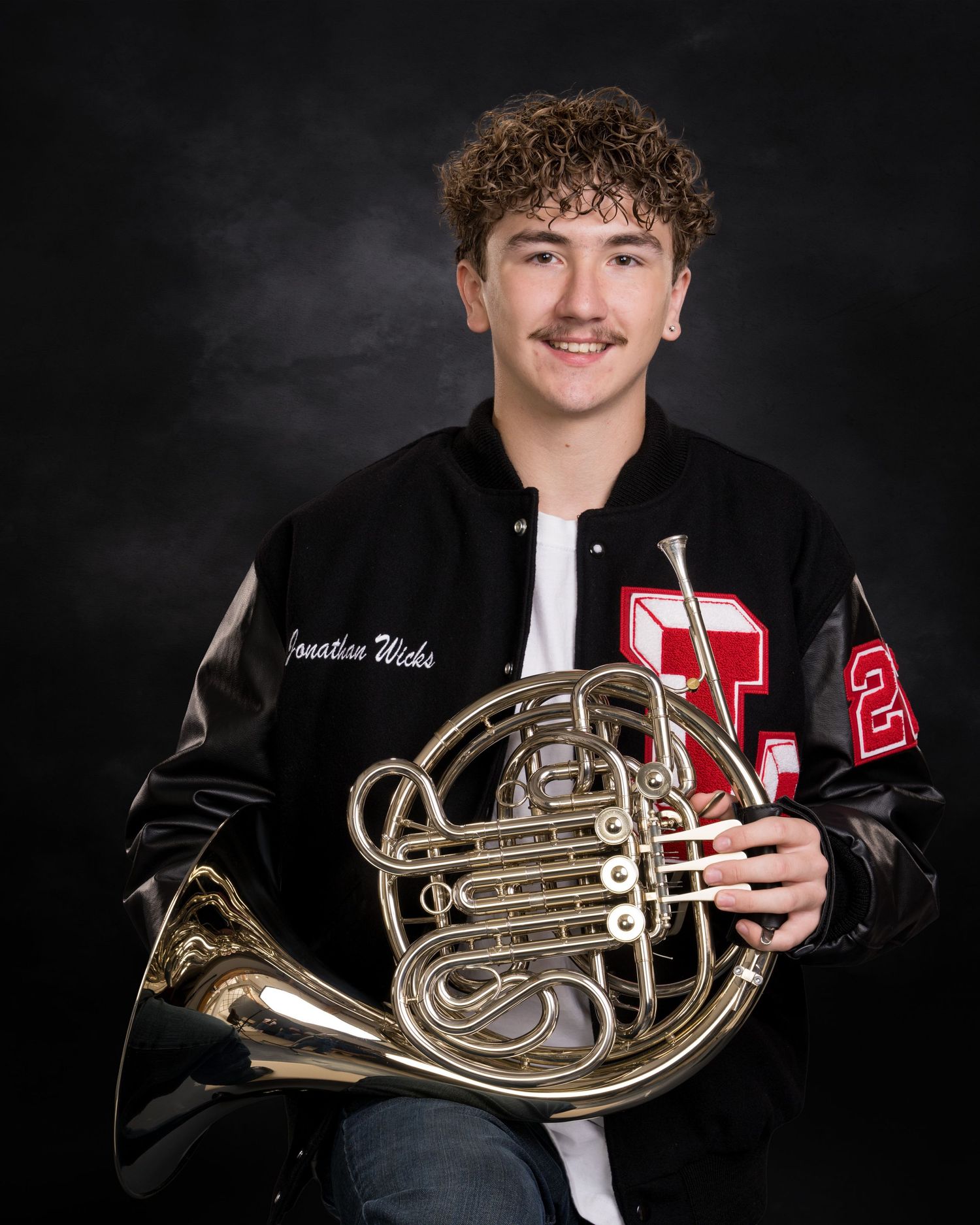 A smiling young man in a varsity jacket holds a French horn while posing against a dark studio background.