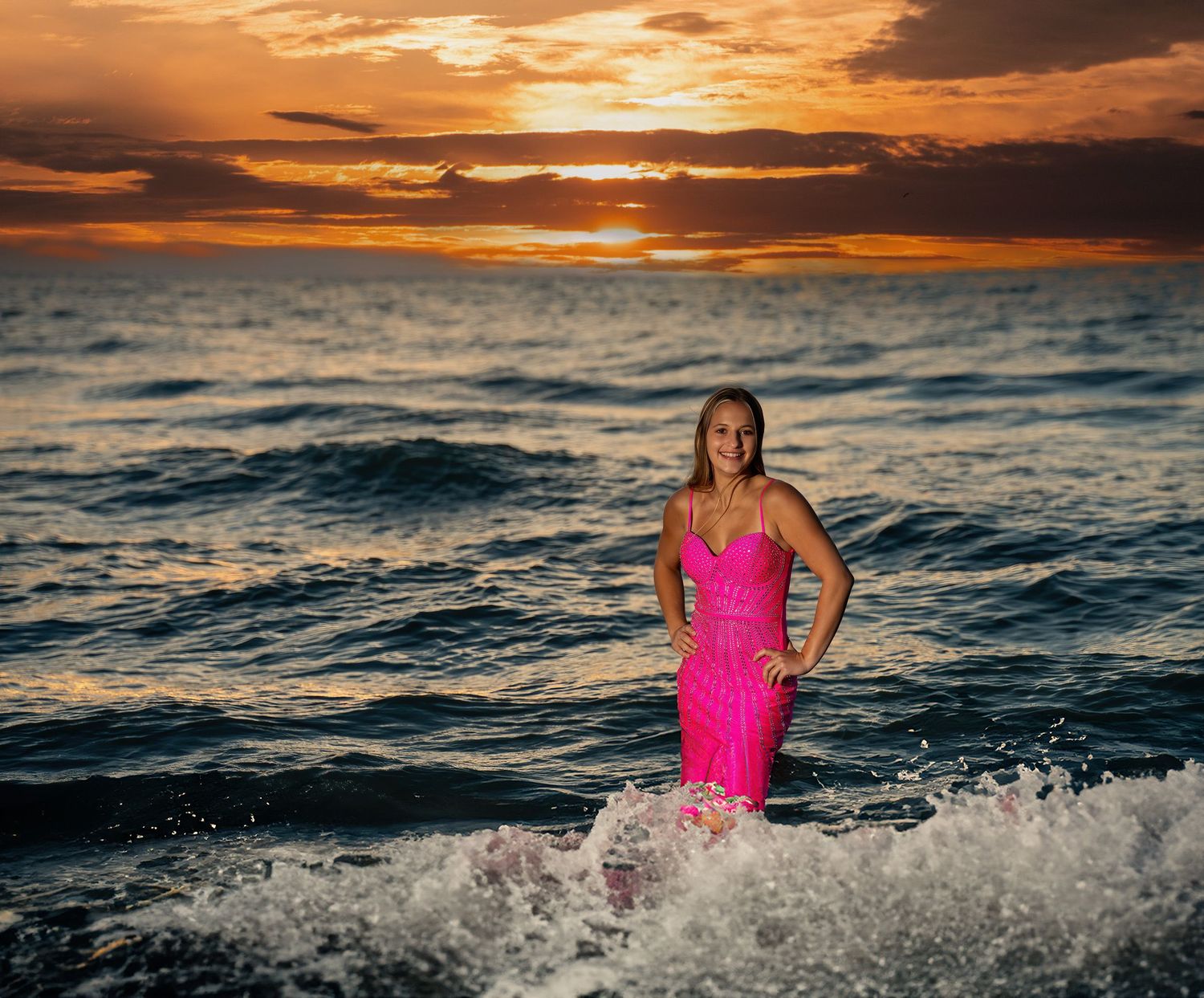 Young woman in pink dress standing in ocean waves during a stunning colorful sunset.