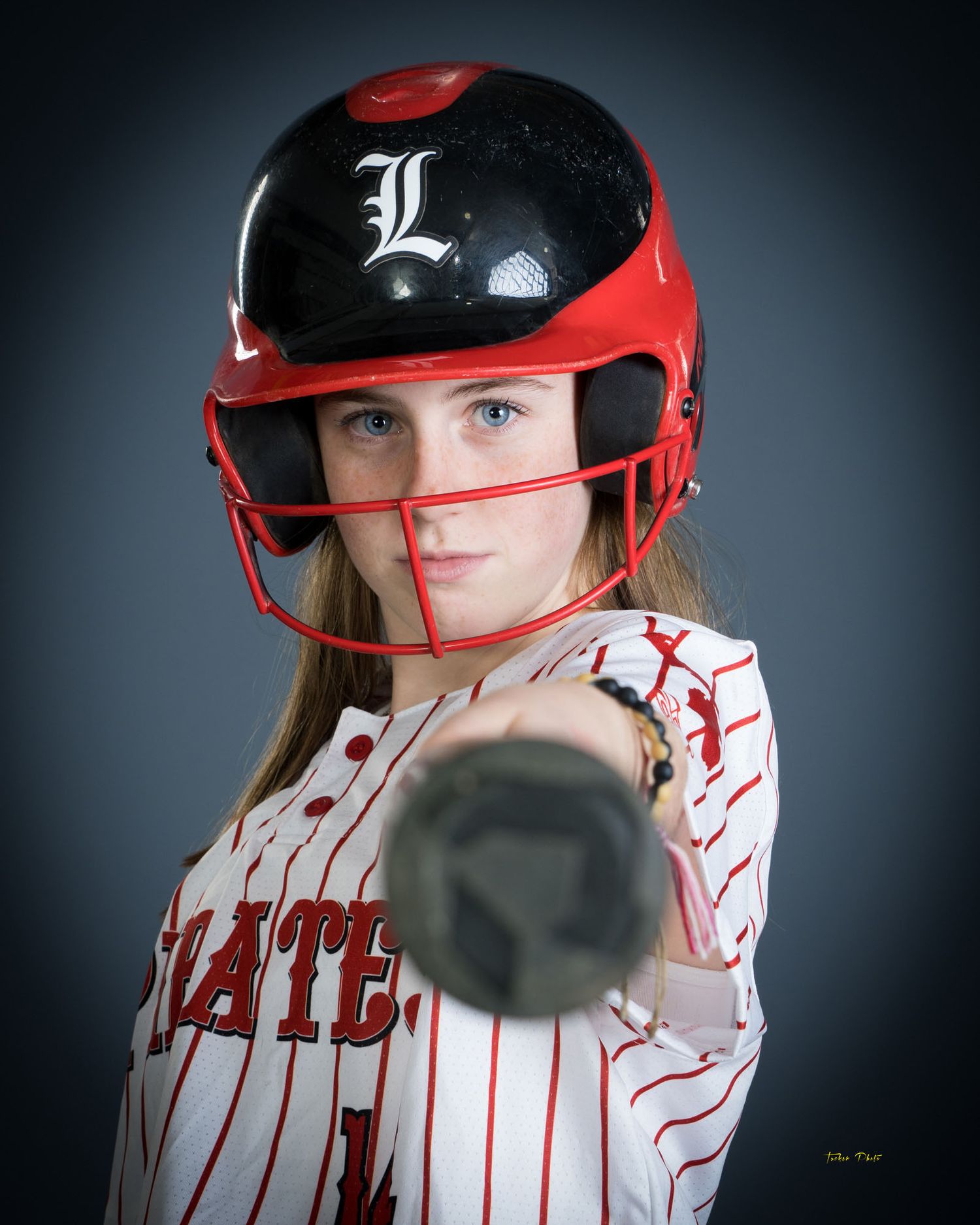 Female softball player in Pirates pinstripe uniform with red helmet pointing bat at camera.