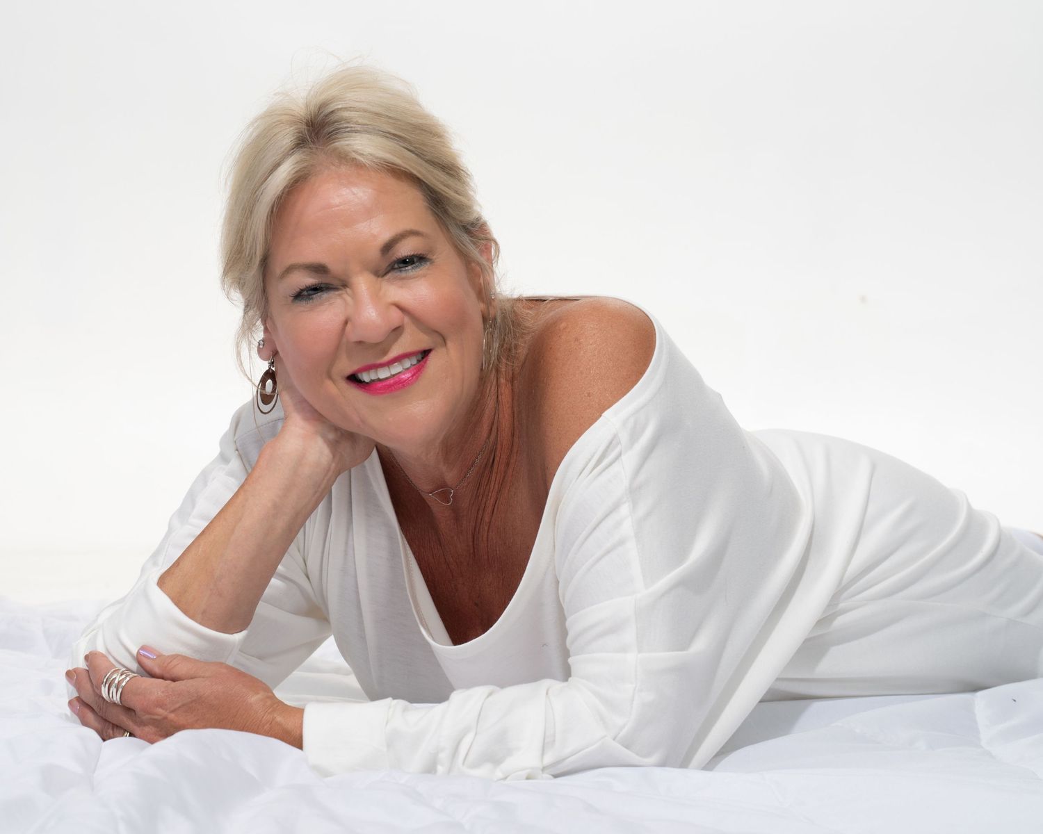 Cheerful blonde woman in white off-shoulder top lying on white bedding with bright smile.