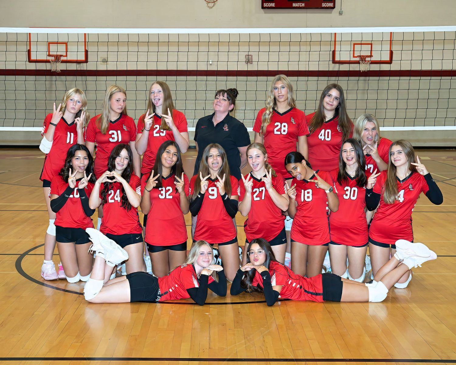 Girls volleyball team in red uniforms posing for group photo in gymnasium with coach.