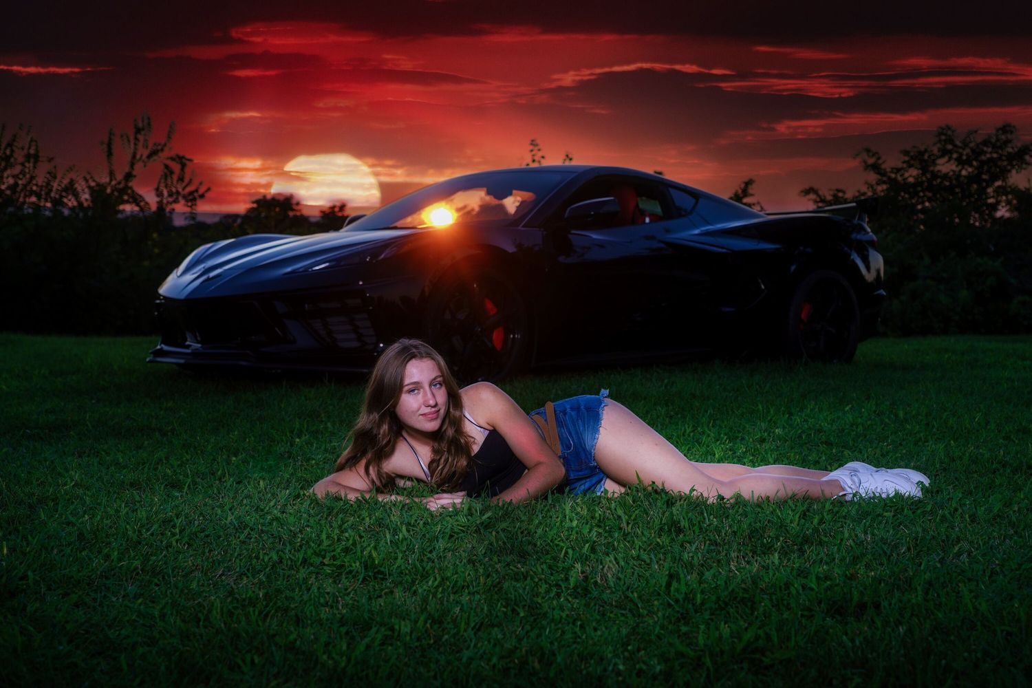 Young woman lying on grass in front of sports car at dramatic sunset.