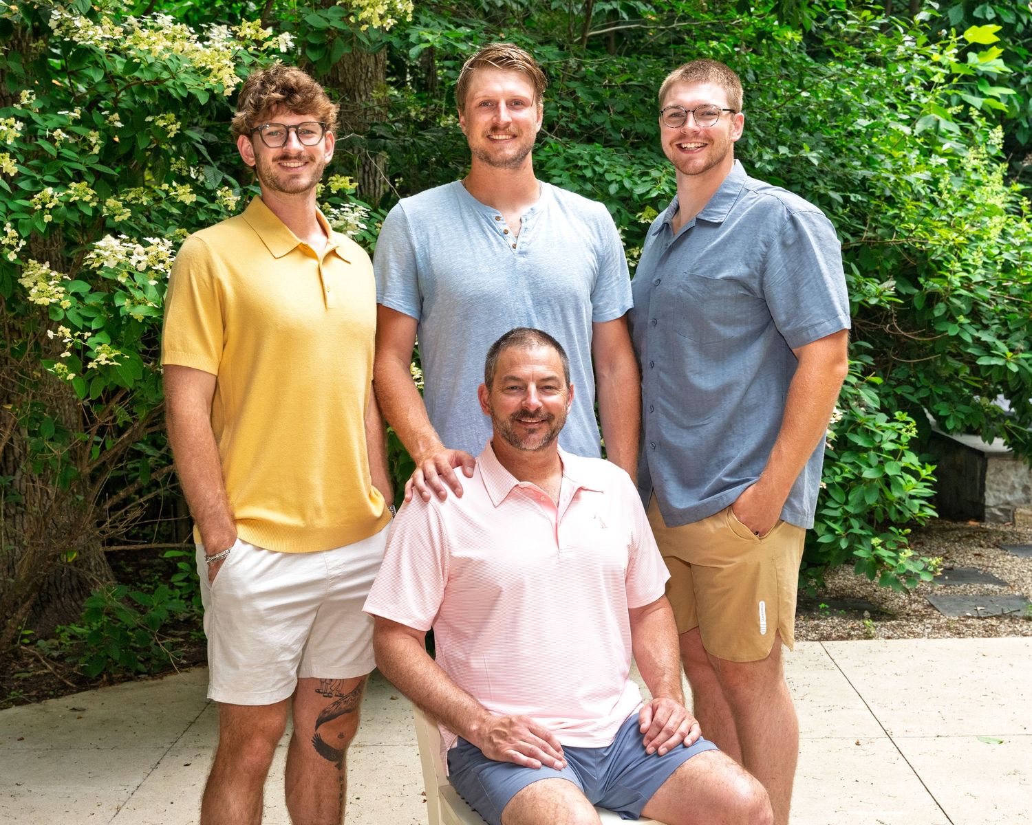 Father posing with three adult sons in colorful polo shirts in outdoor garden setting.