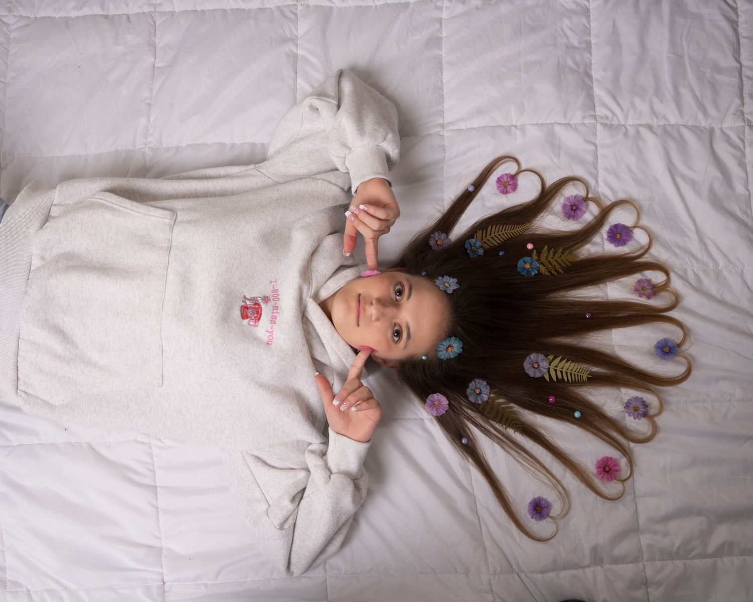 Young girl in white hoodie lies on bed with long hair spread out decorated with colorful flower clips.