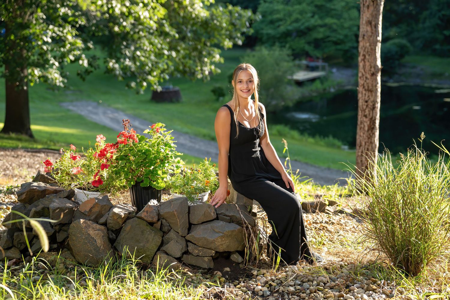 A tall blonde woman in a black maxi dress sits on a stone garden wall surrounded by red flowers and lush greenery.