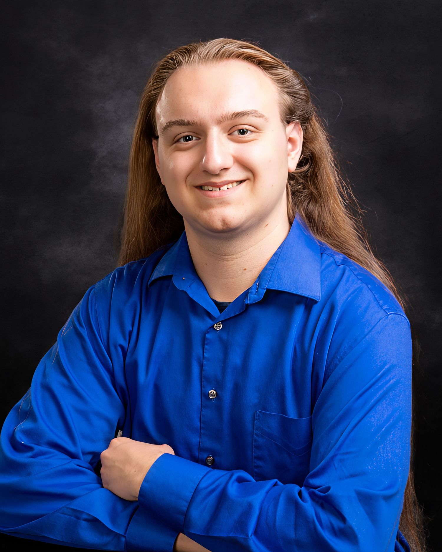 Young man with long hair in blue dress shirt smiles with arms crossed for portrait photo.