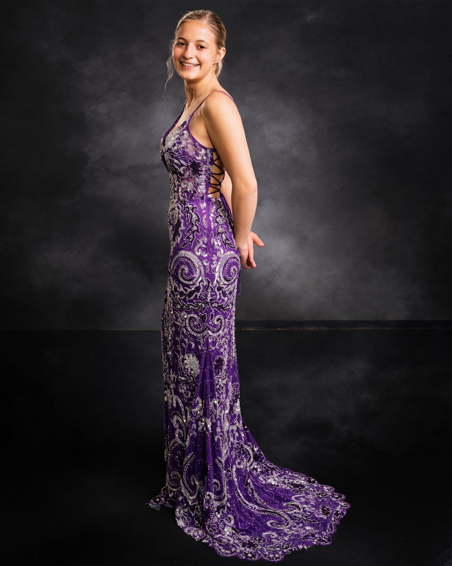 Young woman posing in a purple patterned formal gown with a train on a dark backdrop.