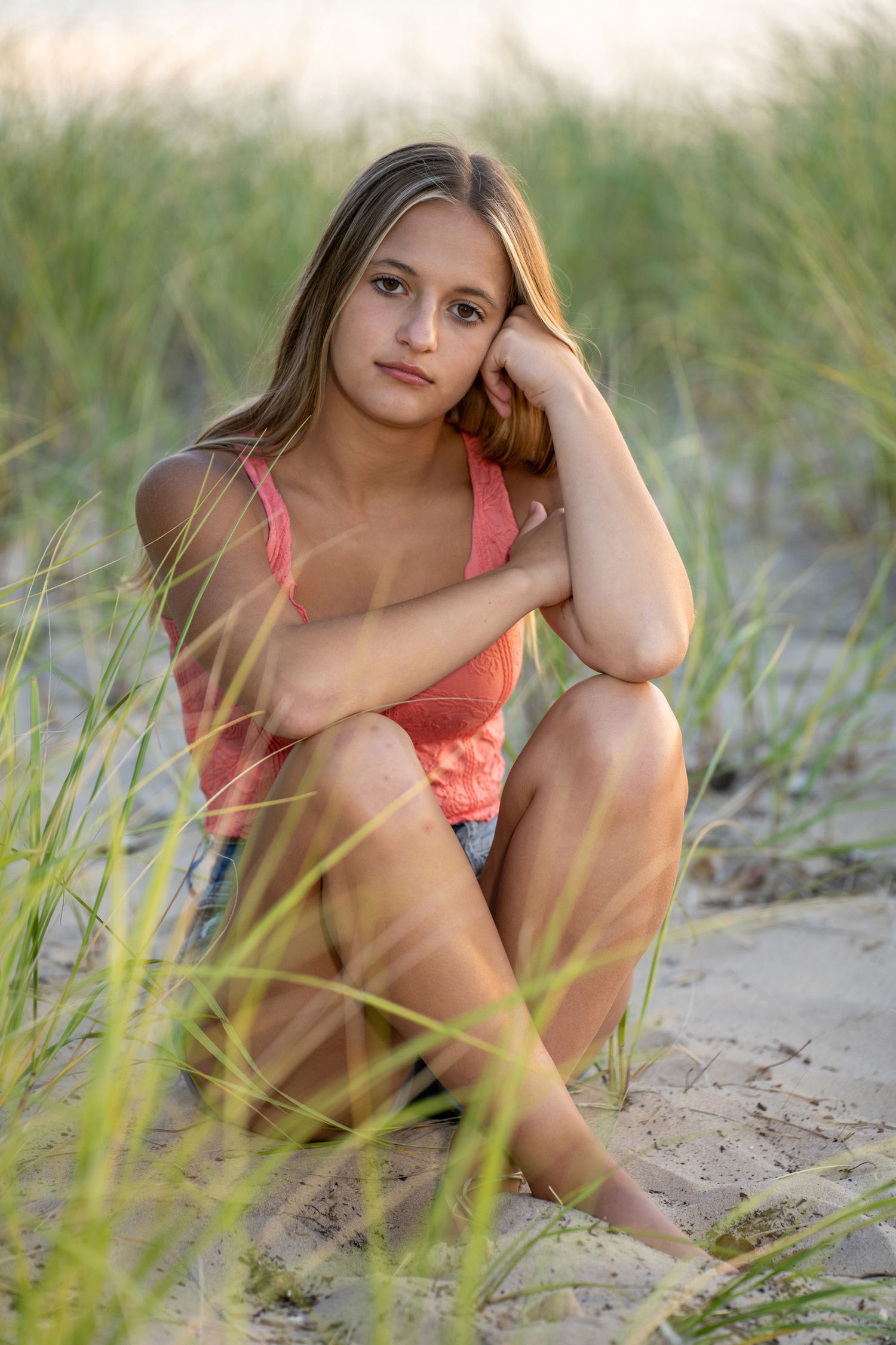 Teen girl in pink tank top sitting on sandy beach dunes surrounded by tall grass at sunset.